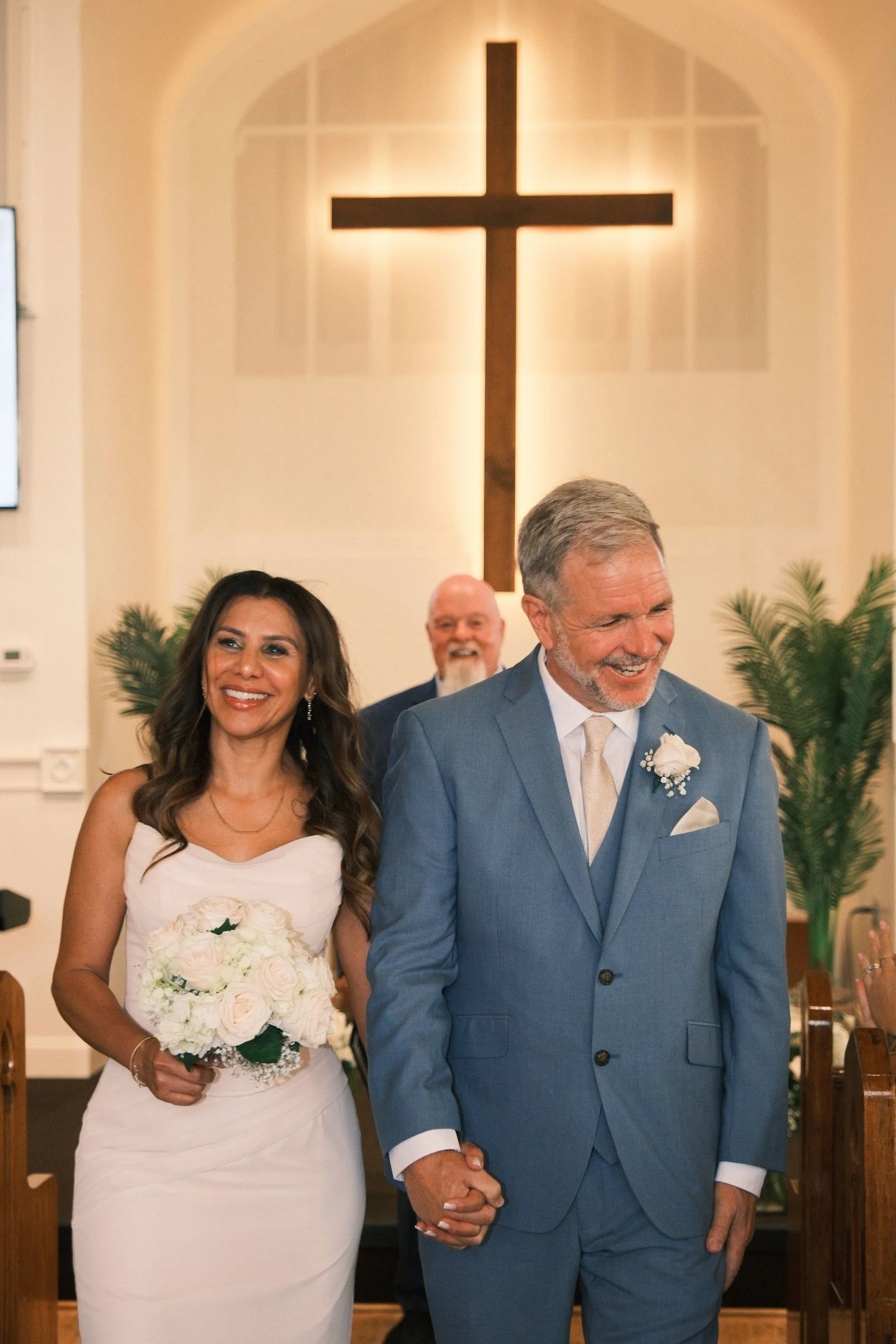 A wedding scene inside a church with a bride and groom holding hands, smiling. The bride has long, dark hair, wearing a strapless white wedding dress, holding a bouquet of white roses. The groom has short grey hair, wearing a blue suit and white tie.