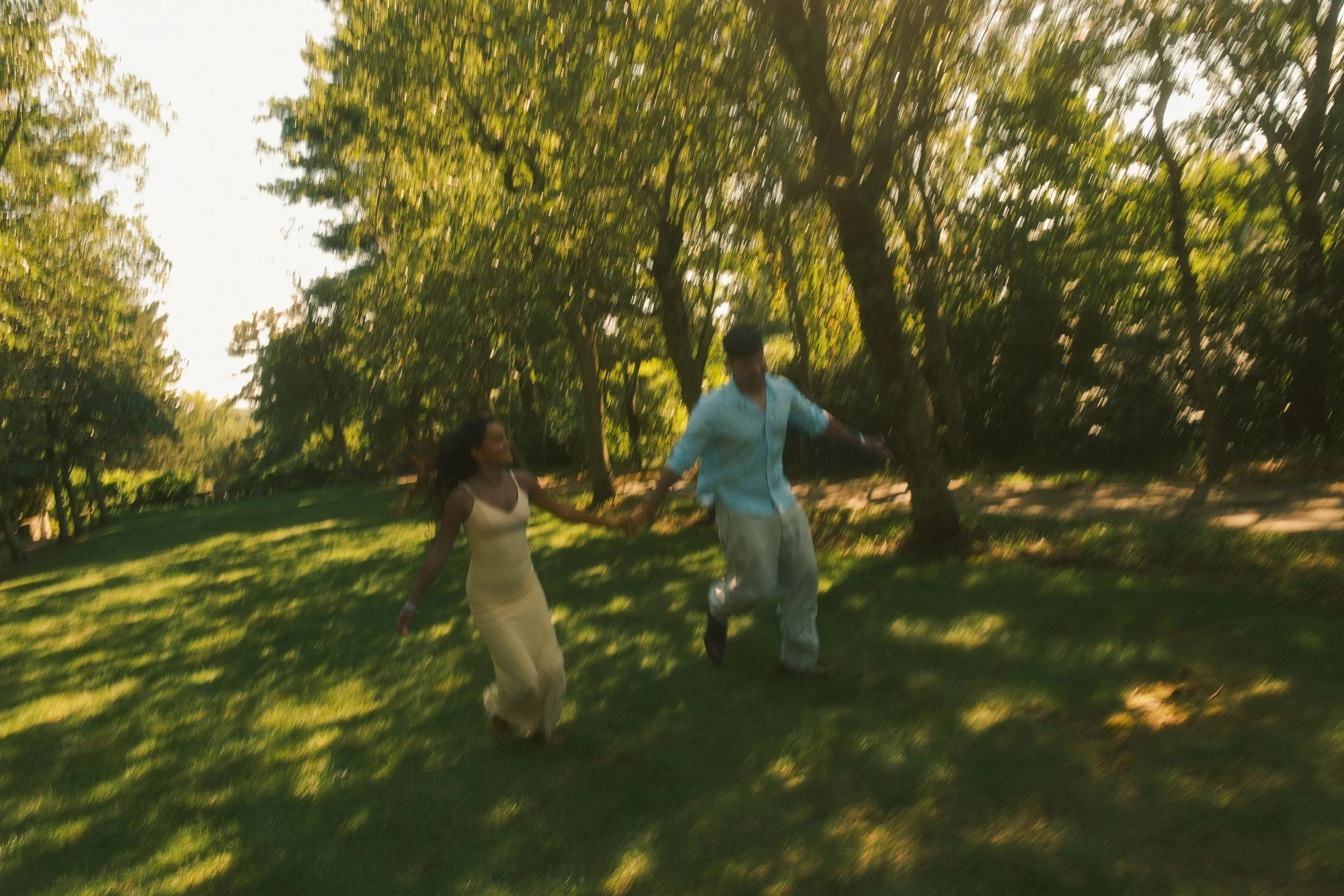 A man and woman dancing outdoors in a park with trees, holding hands and smiling.