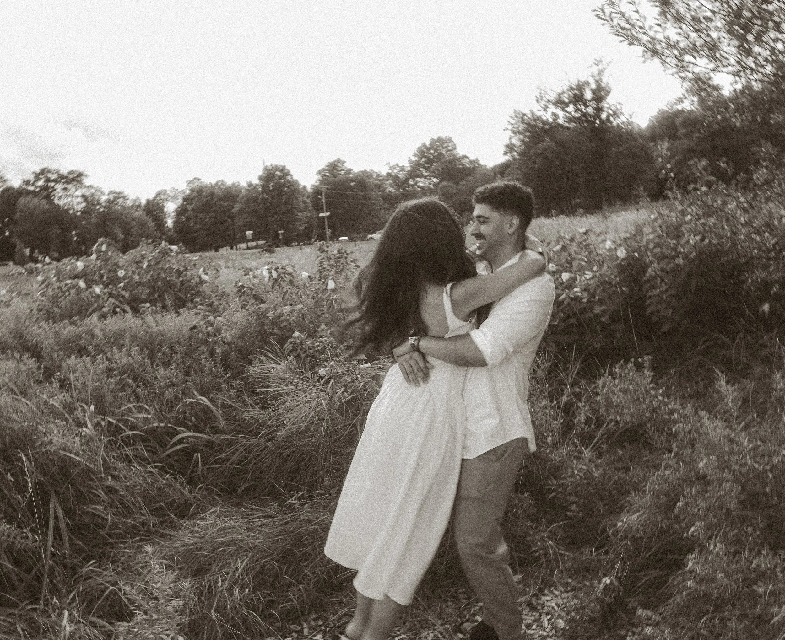 A couple embracing outdoors in a natural setting, with trees and bushes in the background.