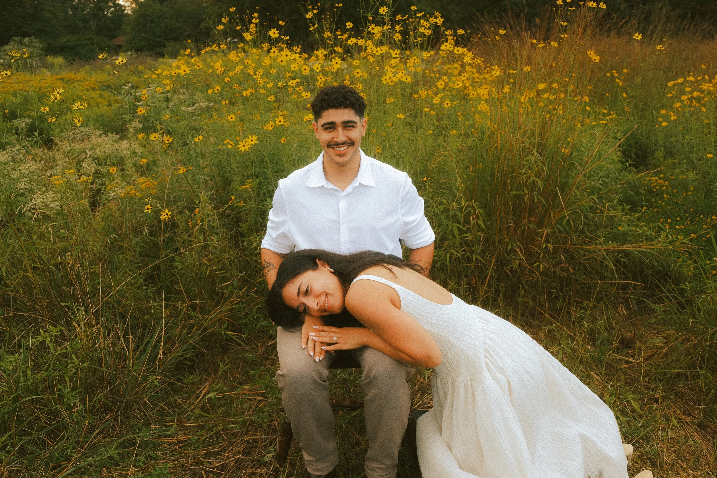 A man with curly hair and a mustache, wearing a white shirt, sits behind a woman with long dark hair and a white dress, who is lying on the ground with her head resting on his knee, in a field of yellow flowers during daytime.