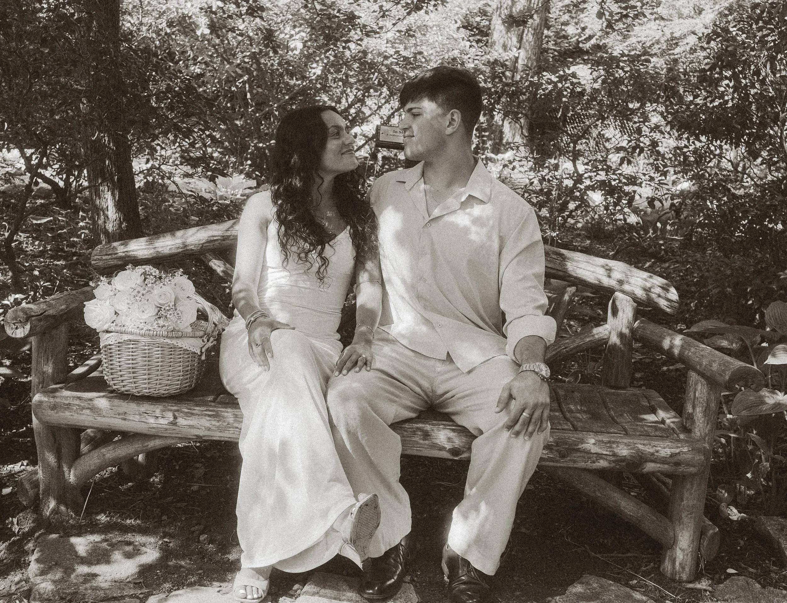 A black-and-white photo of a couple sitting on a wooden bench outdoors, looking at each other affectionately, surrounded by trees and foliage. There is a wicker basket with flowers on the bench.