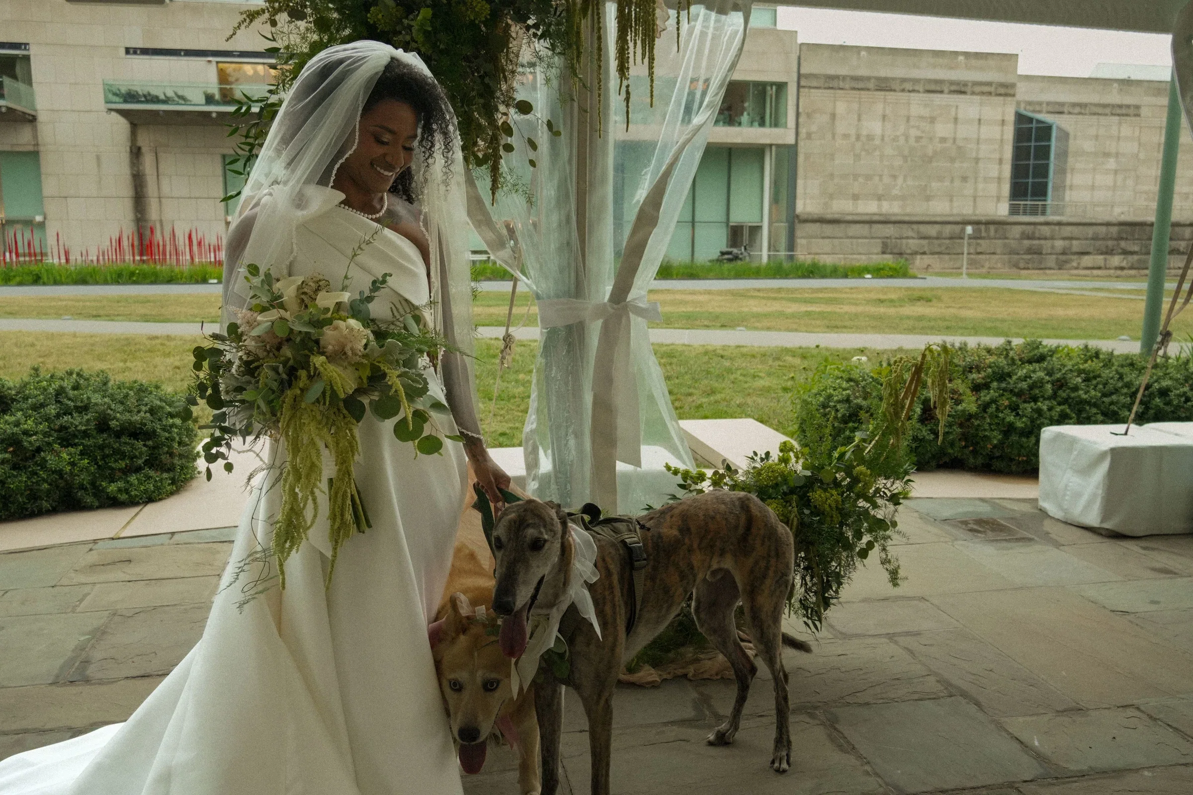 A woman in a wedding dress holding flowers and smiling, accompanied by two dogs, outside a modern building with greenery and glass windows.