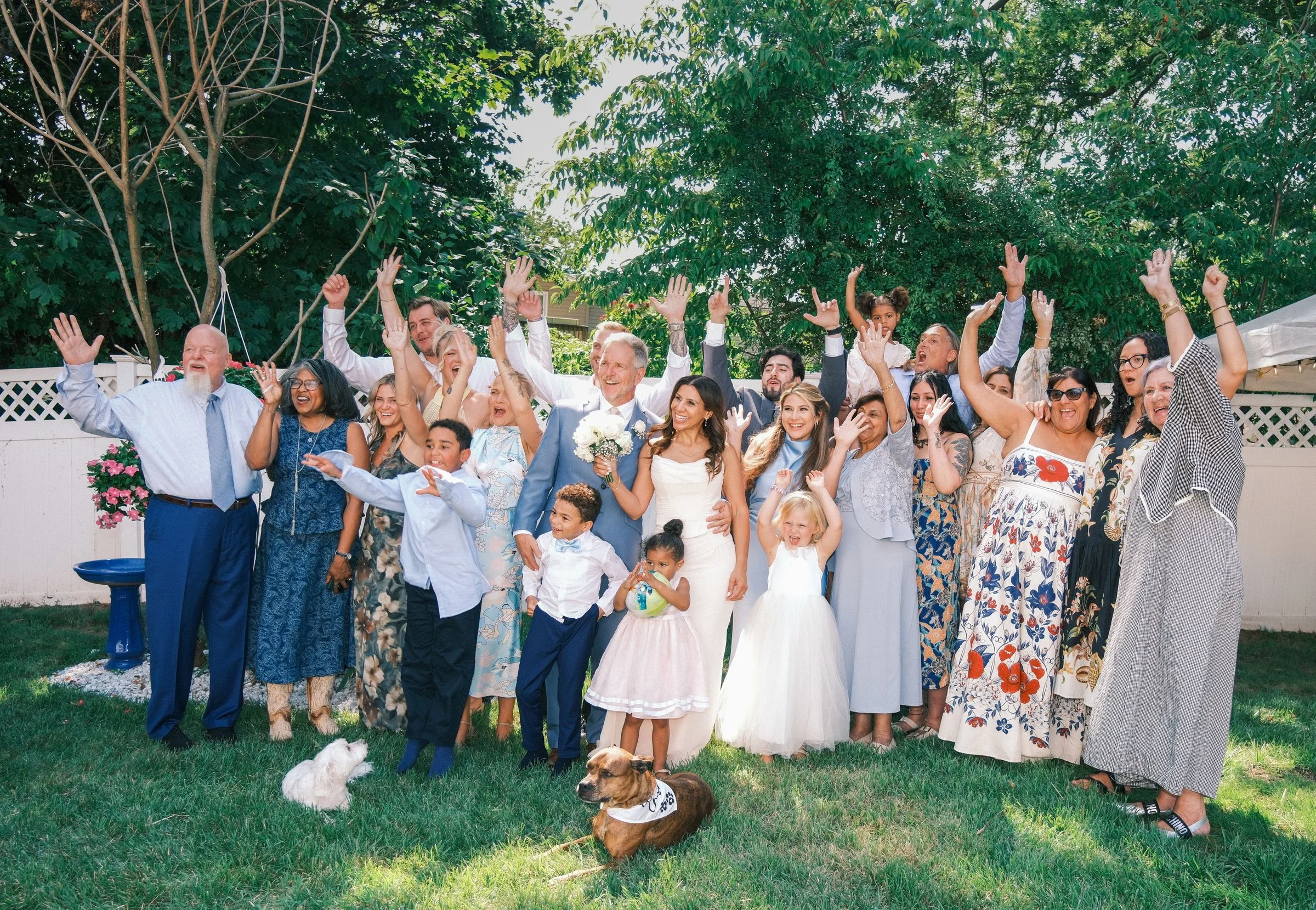 A large group of people, including children, gathered outdoors for a celebration, with many raising their hands and smiling. There are trees and a white fence in the background, and dogs are present on the grass in front of the group.