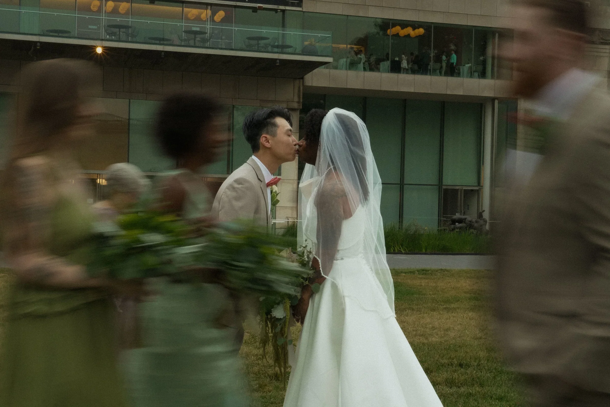 A couple in wedding attire, the man kissing the woman, who is wearing a wedding dress and veil, outside modern glass and concrete building with onlookers in background.