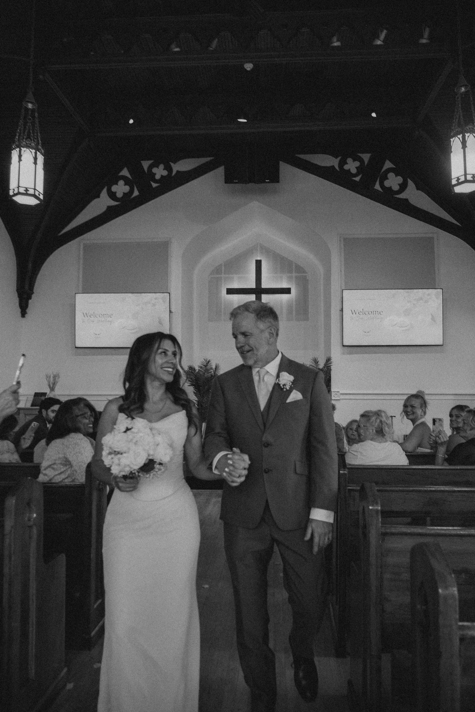A bride and groom are standing together inside a church, holding hands and smiling. The bride is in a white wedding dress holding a bouquet, and the groom is in a dark suit with a boutonnière. The background features a cross on the wall and wedding g