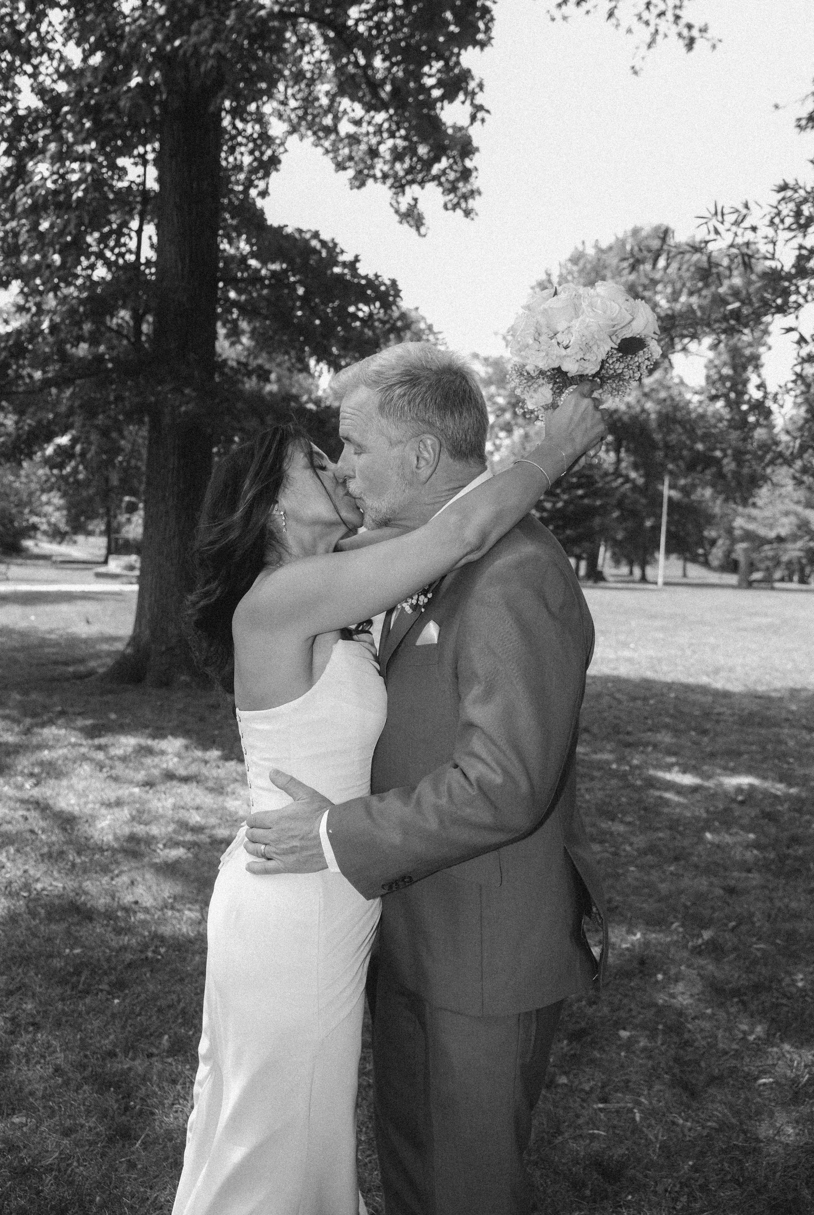 A black and white photo of a couple kissing outdoors. The woman is in a white dress and the man is in a suit. The woman is holding a bouquet of flowers. They are standing near large trees, with grass and park scenery in the background.