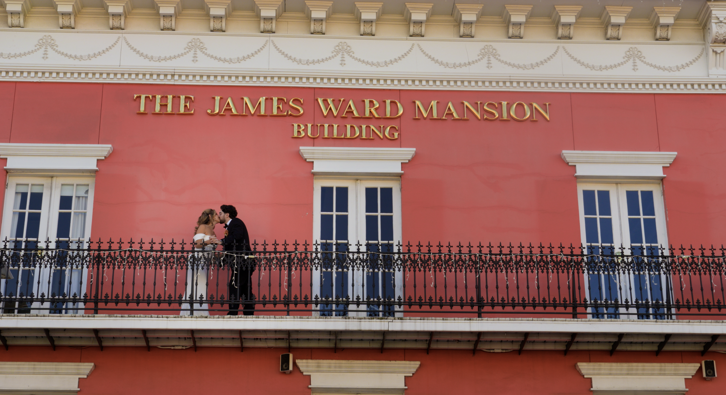 A couple in wedding attire stands on a balcony in front of the James Ward Mansion building, which has a pink exterior and golden lettering.