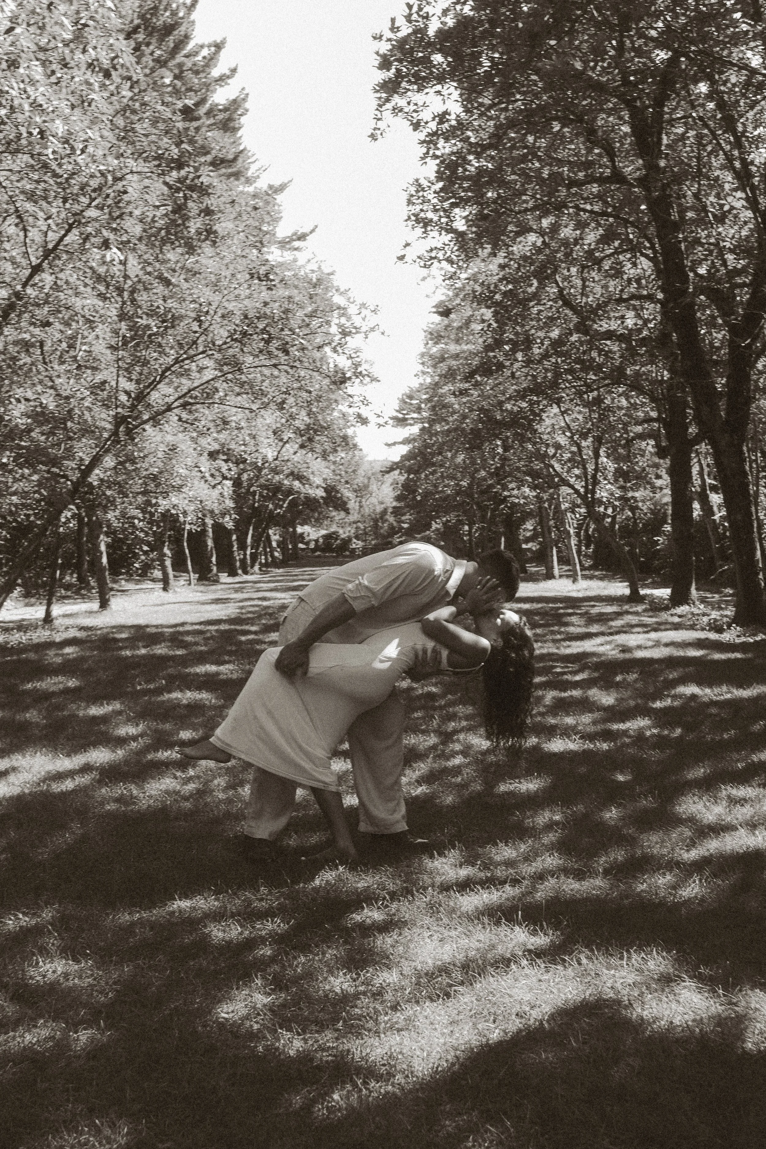 Two people practicing acro yoga outdoors in a park with trees, one person holding the other in a backbend pose, both wearing casual clothing.