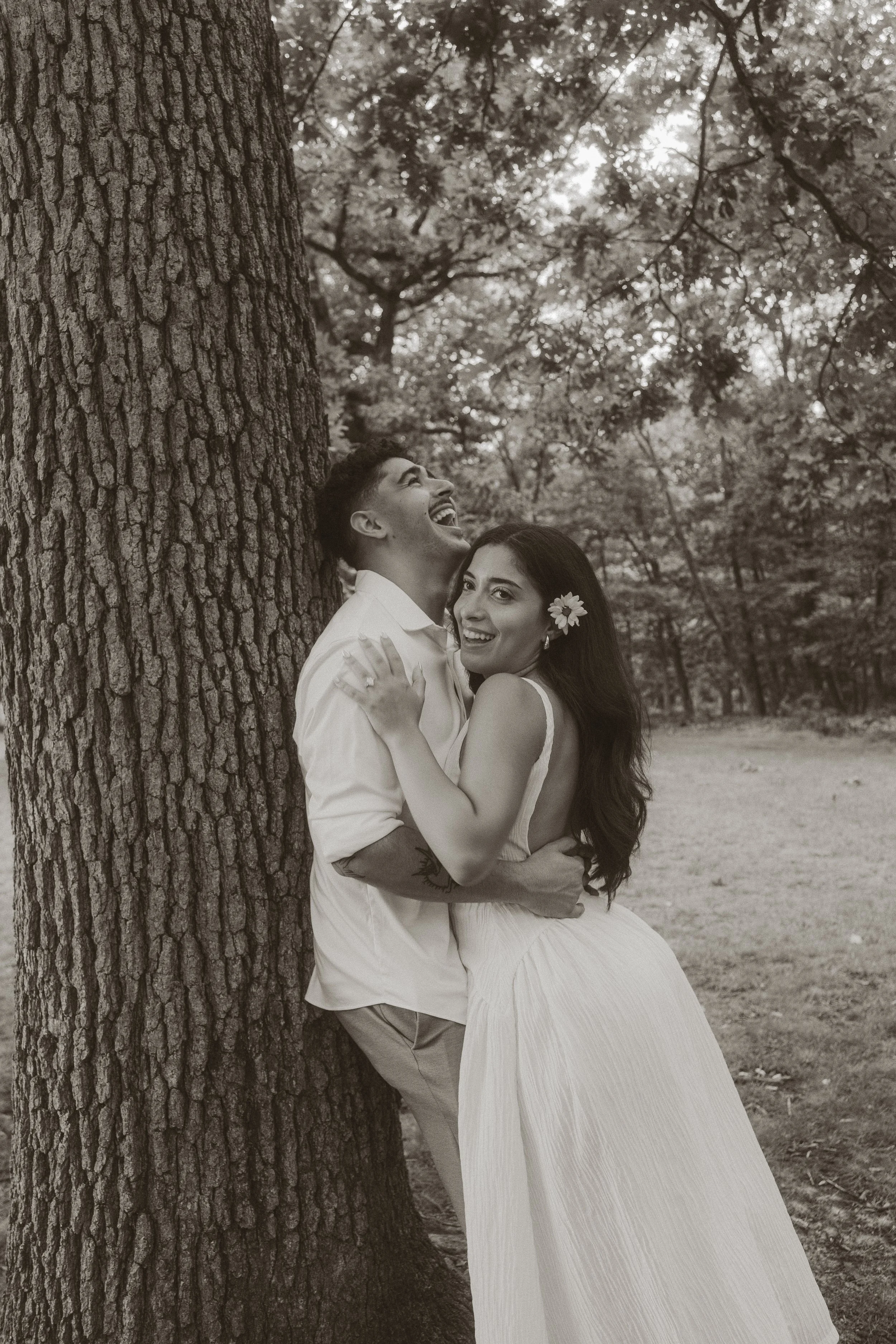 A happy couple in a park, leaning against a large tree, embracing and smiling, with the woman wearing a flower in her hair.