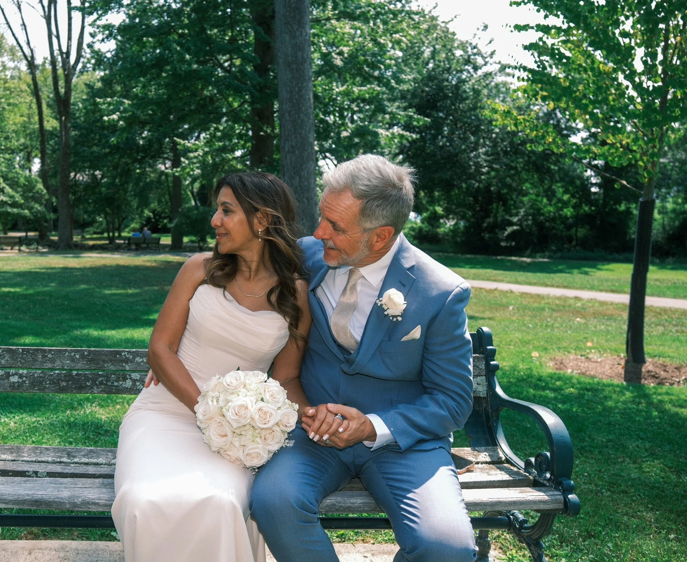 A bride and groom sitting on a park bench surrounded by green trees during the daytime. The bride is holding a bouquet of white roses, and the groom is dressed in a light blue suit with a white shirt and tie.
