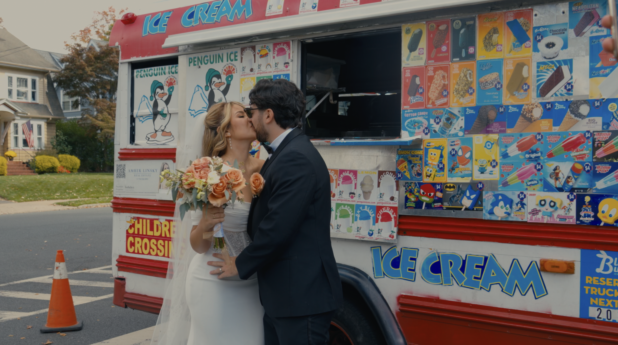 A newlywed couple sharing a kiss in front of an ice cream truck on a residential street. The bride is holding a bouquet of flowers, and the groom is in a black tuxedo. The ice cream truck is decorated with colorful ice cream and frozen treats advertisements.