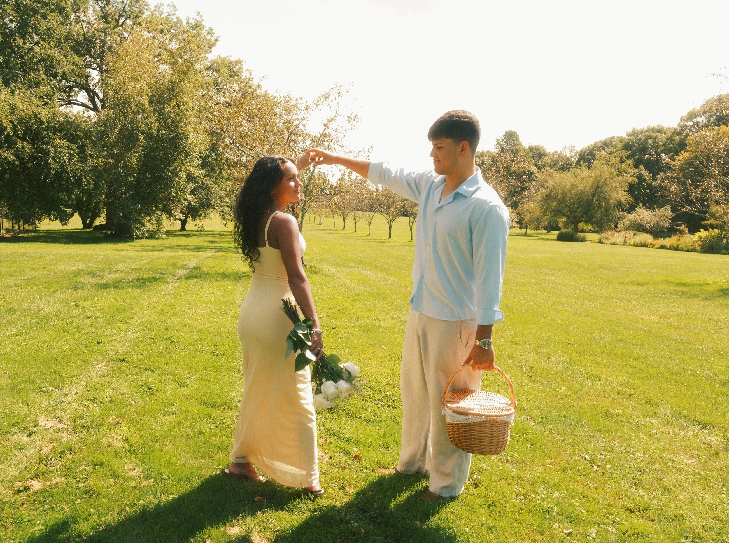 A young man is reaching out to touch the forehead of a young woman in a yellow dress, who is holding a bouquet and a wicker basket, in a park with green grass and trees.