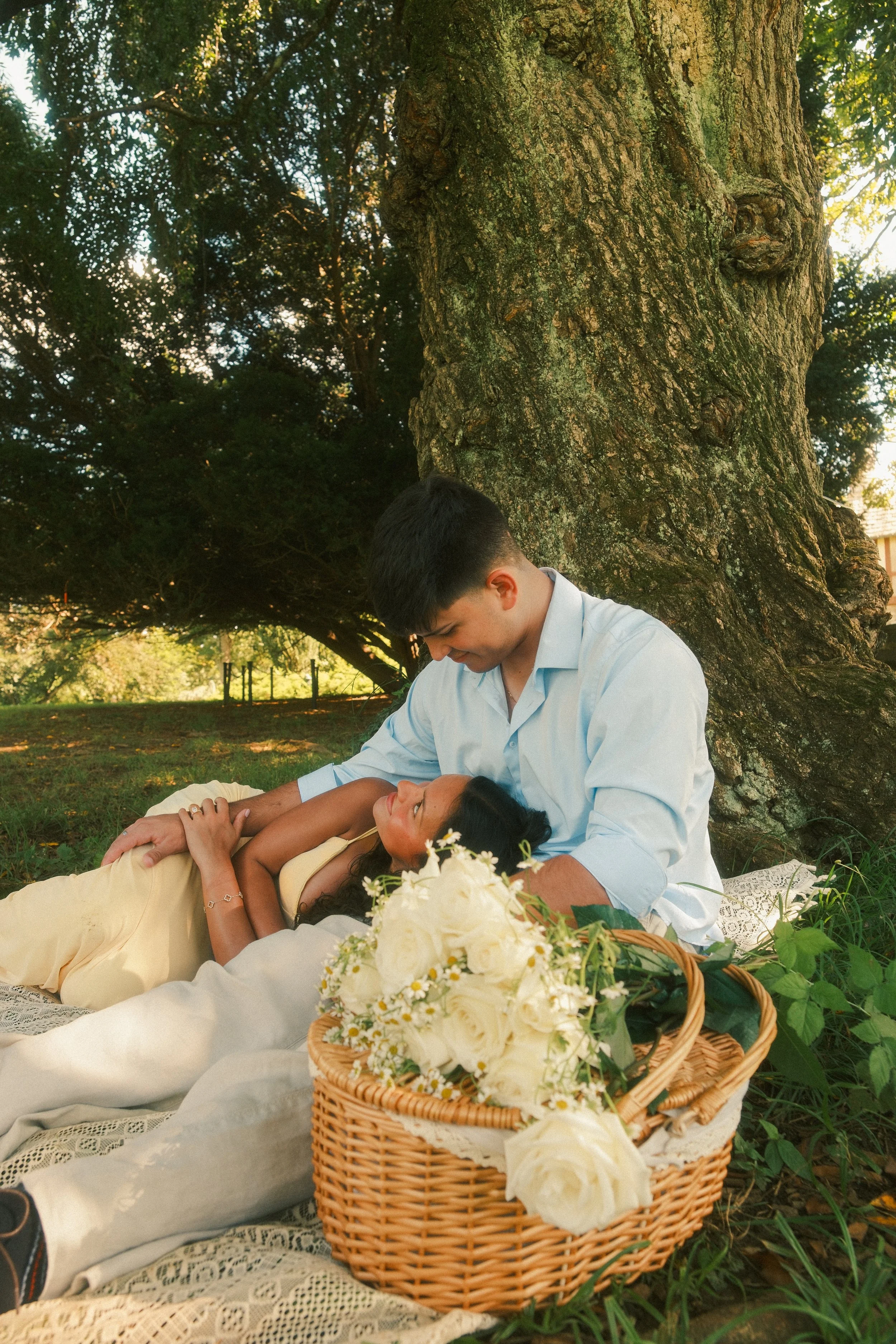 A young man and woman lying under a large tree, with the woman resting her head on the man's lap. There is a basket filled with white roses and daisies in front of them on a blanket, during a sunny day in a park.