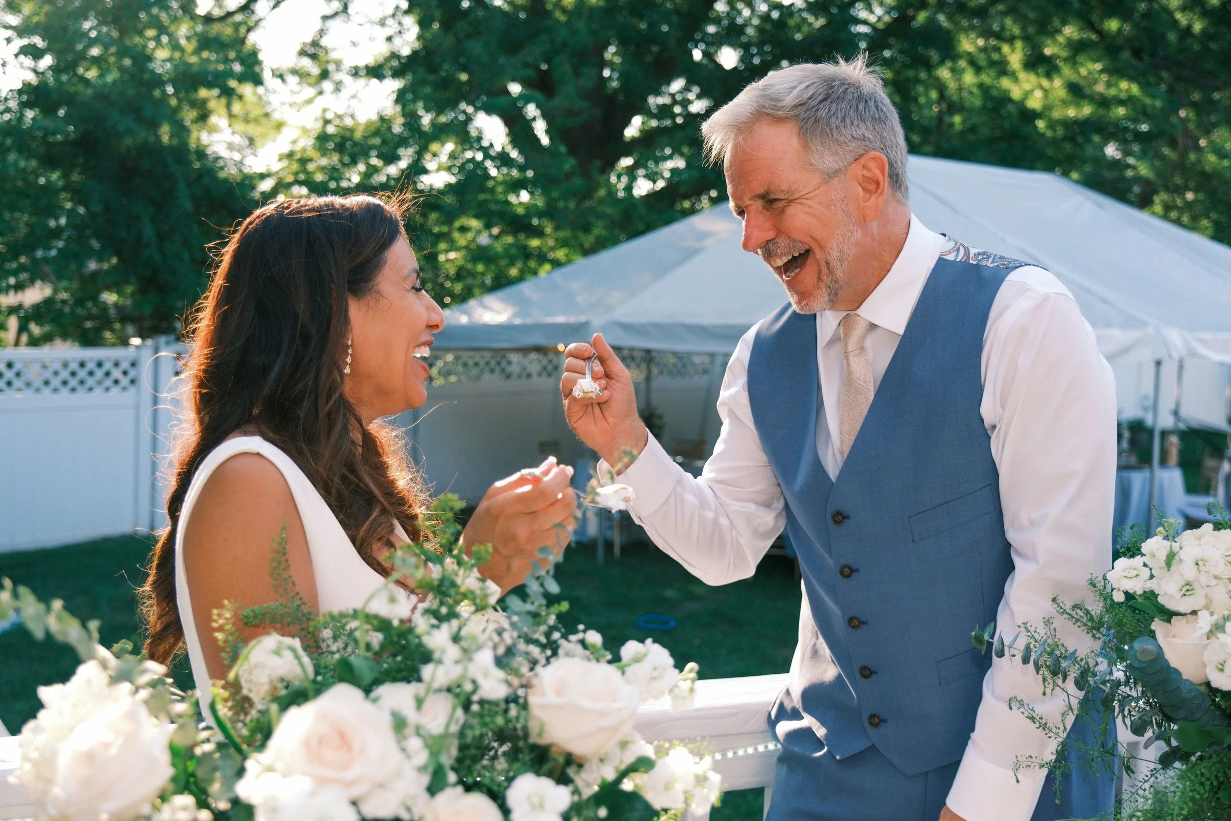 A joyful groom with gray hair and a beard presenting a ring to a smiling bride with long dark hair during a wedding celebration outdoors, surrounded by white flowers and green trees in the background.