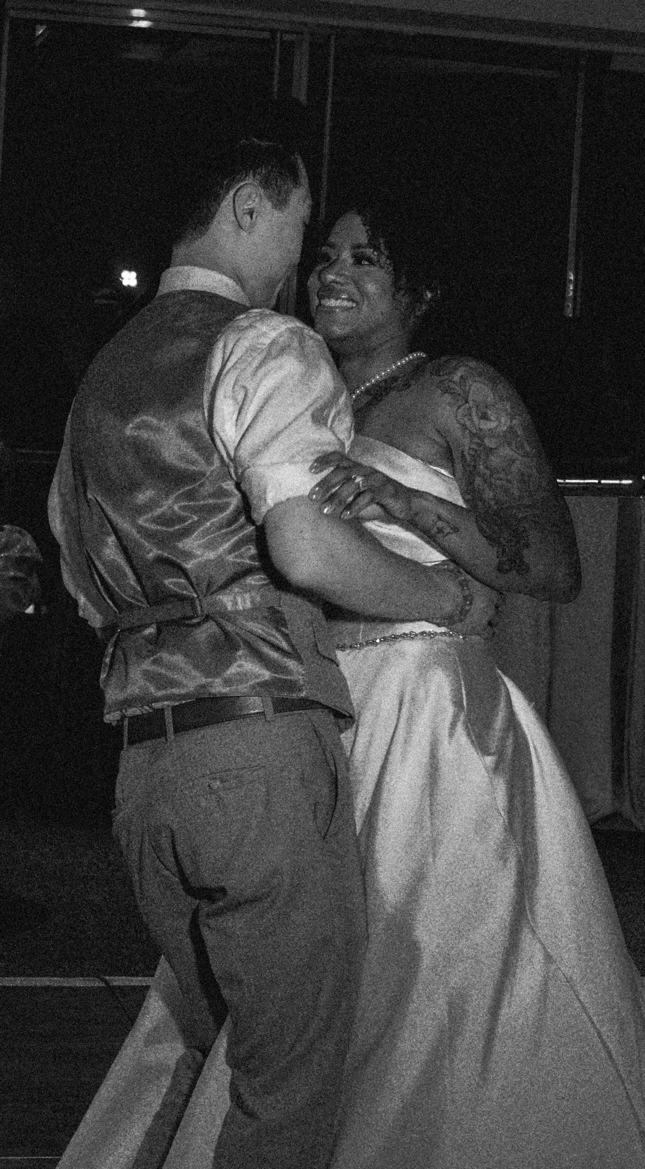A couple dancing closely at a formal event, with the woman smiling and wearing a dress and pearls, and the man in a dress shirt and vest, in a dimly lit room.