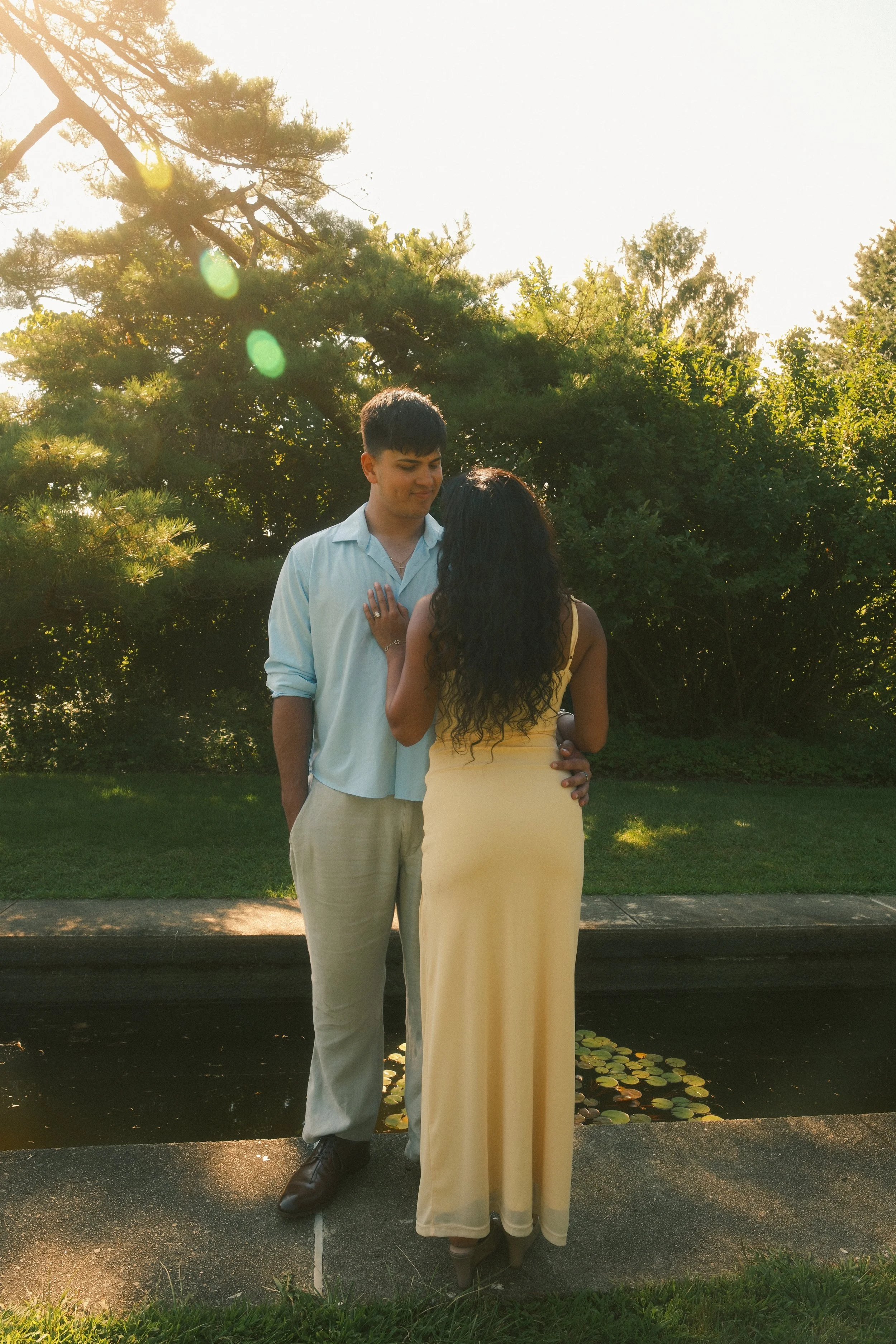 A couple stands by a pond surrounded by lush green trees, with the woman holding the man's chest in a romantic pose, both looking at each other, with sunlight filtering through the leaves.