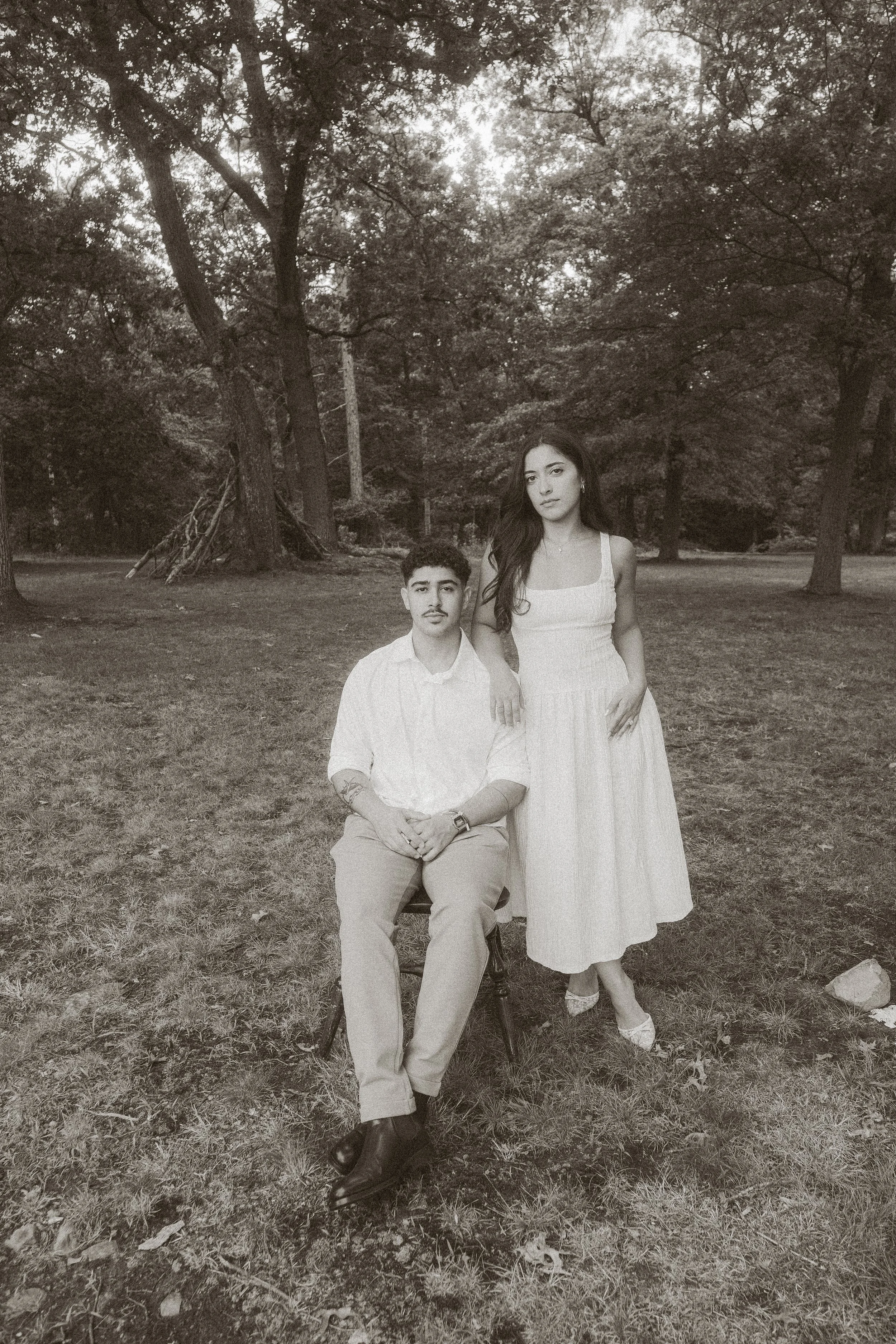 A black and white photo of a man and woman in a park surrounded by trees. The man is sitting on a chair, wearing light-colored pants and a light shirt, with his hands resting on his lap. The woman, standing next to him, is wearing a long, sleeveless 