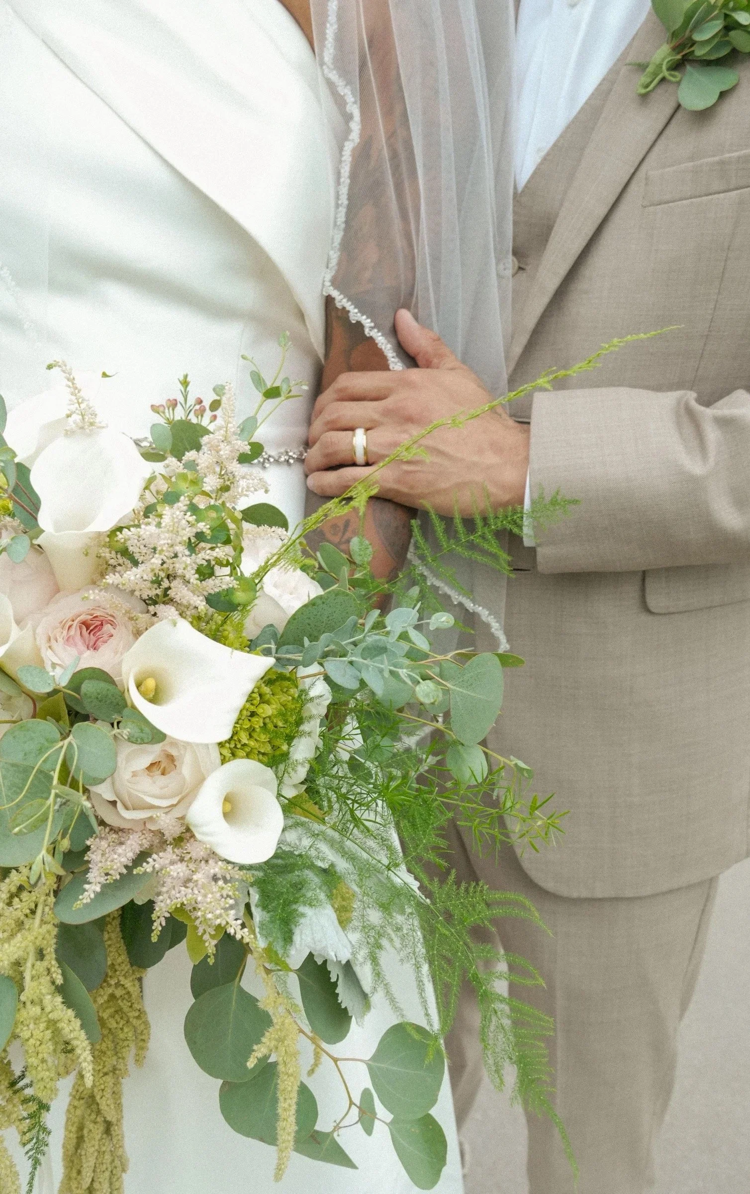 A white wedding dress with veil, a bouquet of white and blush pink flowers, and a person in a beige suit with a wedding band, standing near a beige sofa and a potted plant.