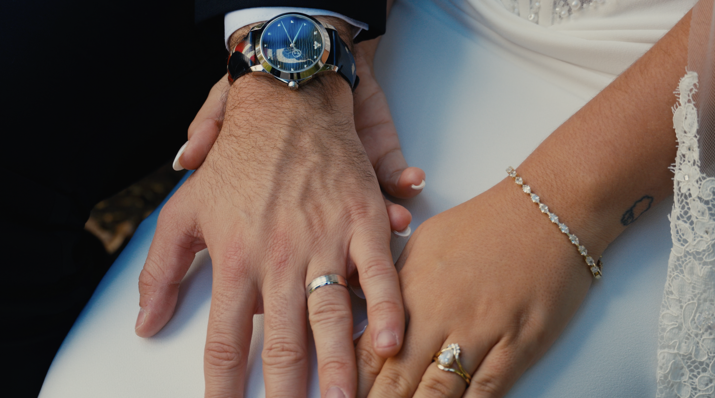Close-up of a couple's hands intertwined during a wedding ceremony, showing wedding rings, watches, and jewelry, with lace fabric visible.