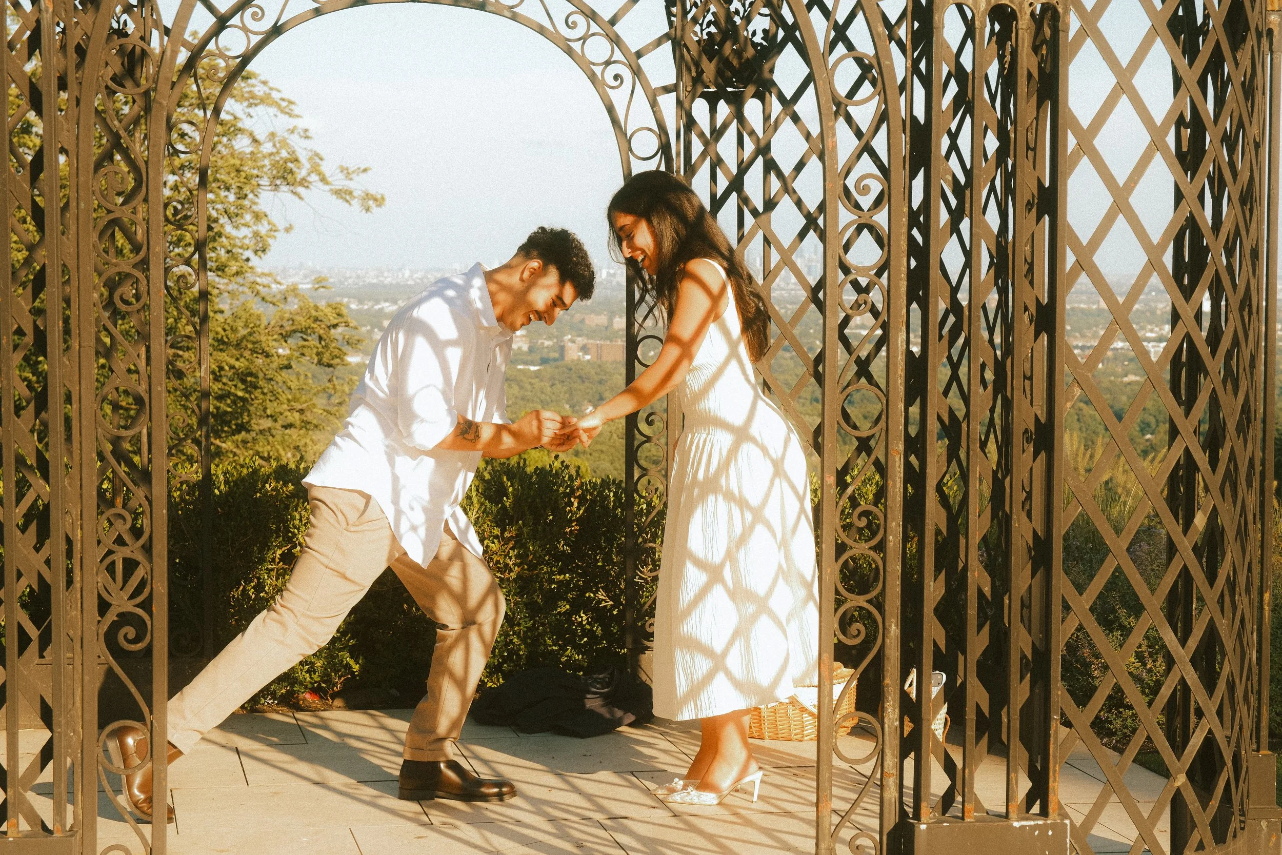 A couple is dancing happily under a decorative metal archway, with the man in a white shirt and beige pants, and the woman in a white dress and high heels, in an outdoor setting with trees and a city skyline in the background.