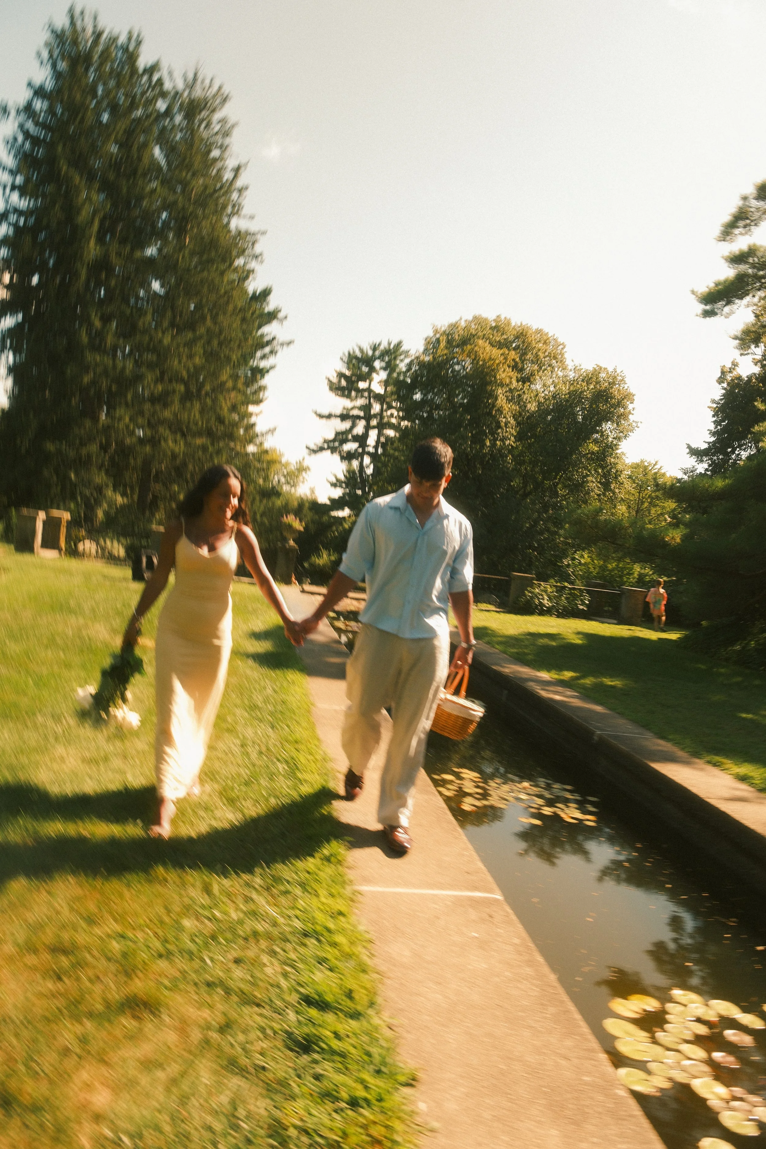 A young man and woman walking hand in hand along a park sidewalk during daytime, with trees and water lilies on the side.