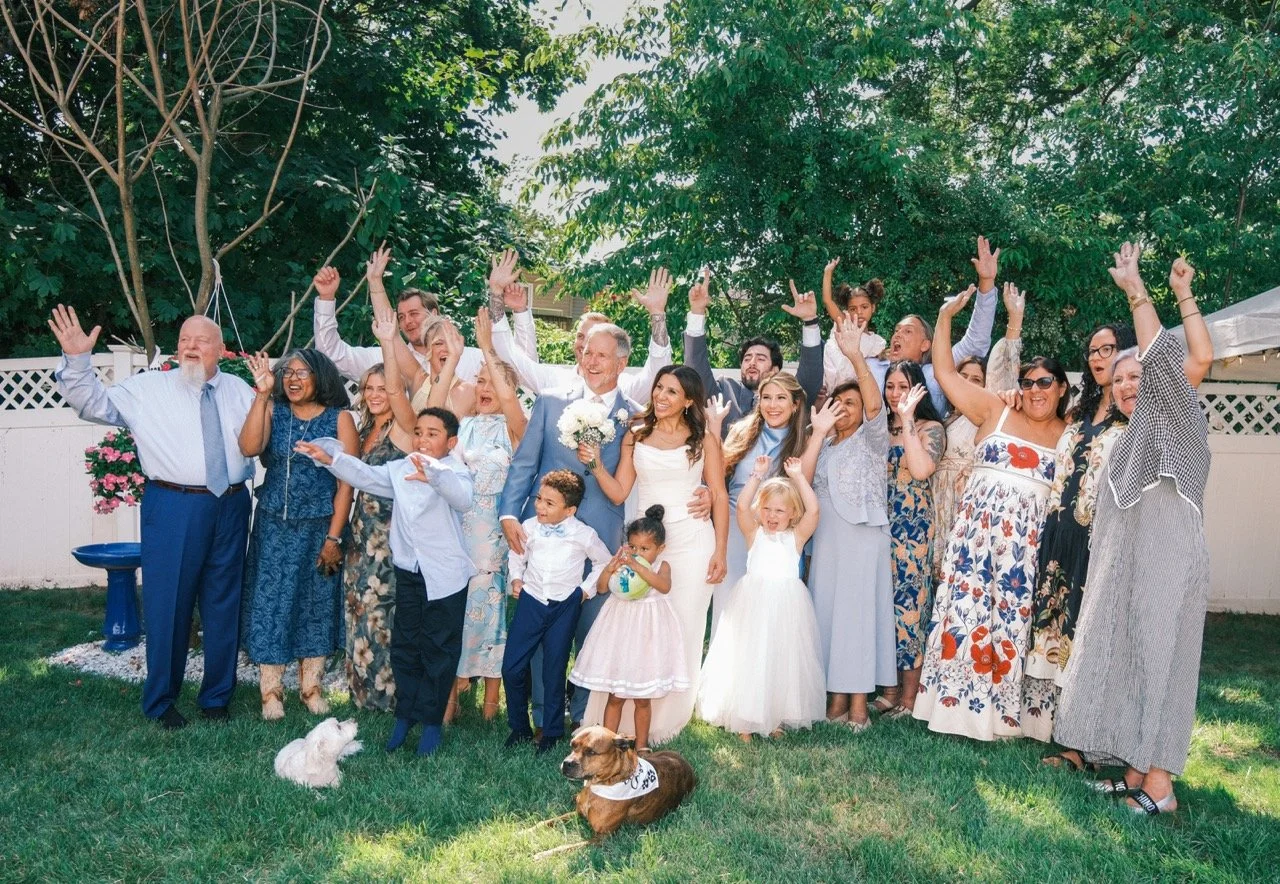 A large group of people, including children and adults, celebrating outdoors on a sunny day, with many raising their hands and smiling. Two women in the center are dressed in a wedding gown and suit, indicating a wedding celebration. There are dogs on the grass in front, and the background features trees and a white fence.