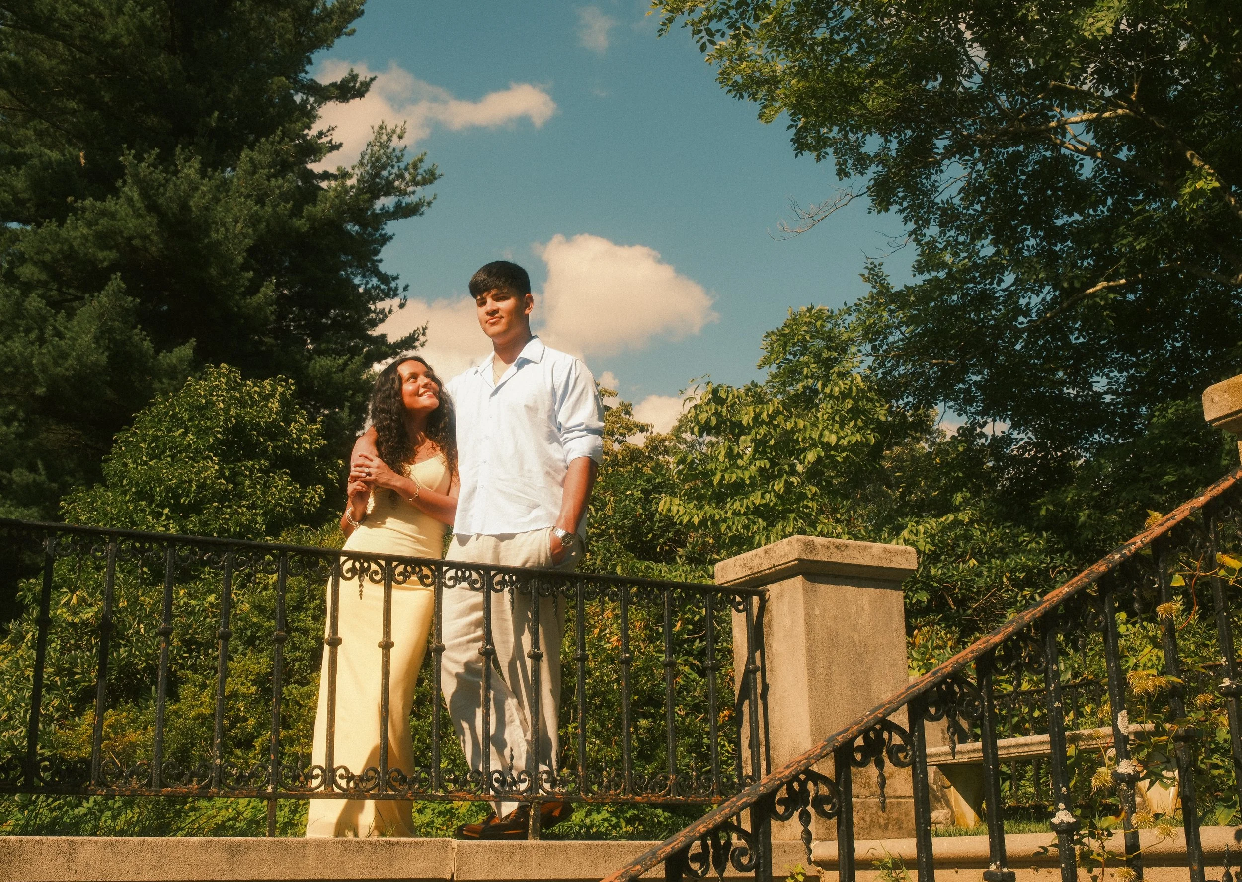 A young couple standing on a stone staircase outdoors surrounded by trees, with the woman looking up at the man as they smile. The sky is blue with a few clouds.