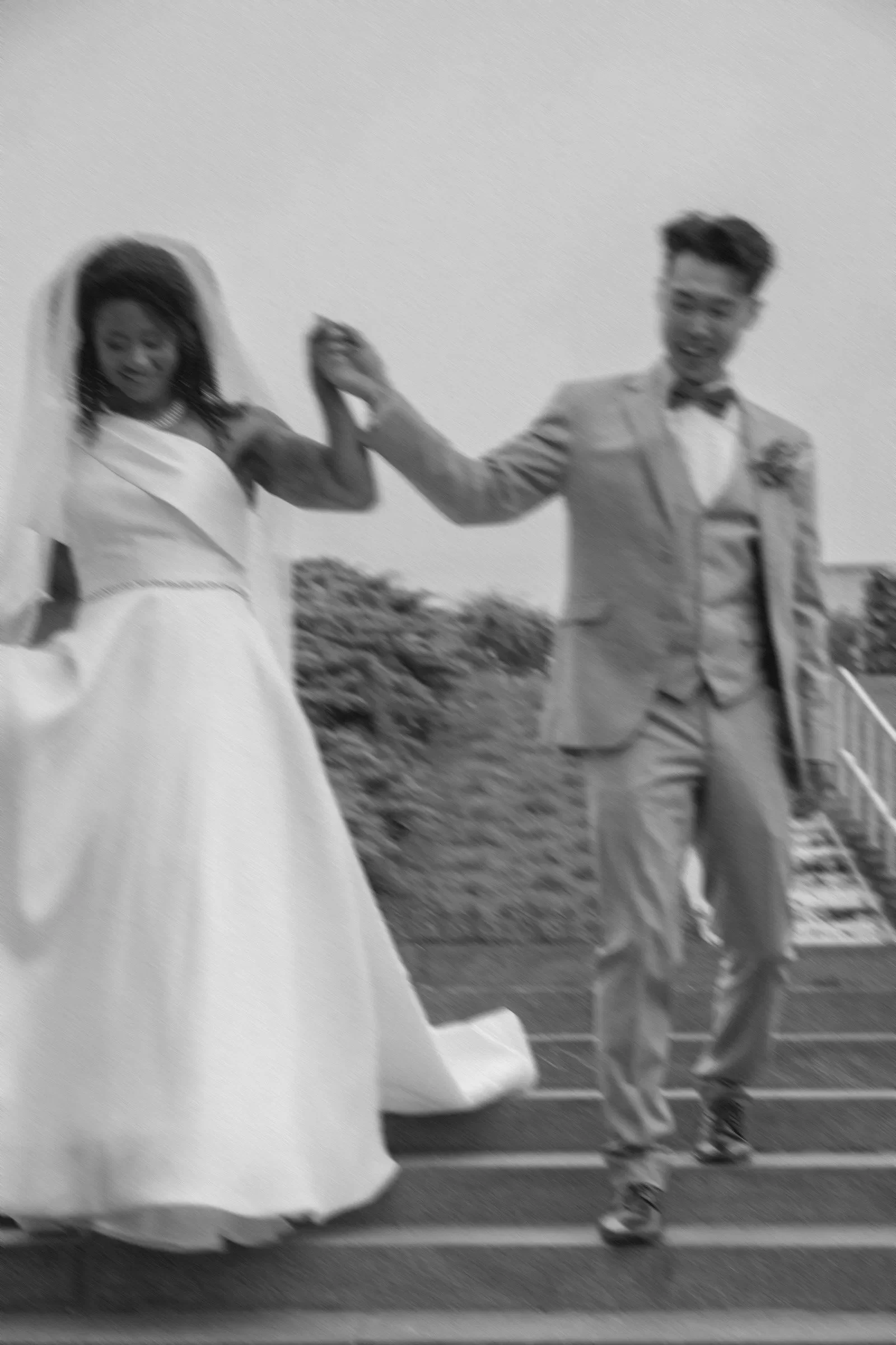 Blurry black and white photo of a newlywed couple, with the bride in a wedding dress and veil and the groom in a tuxedo, holding hands and dancing or celebrating outdoors.