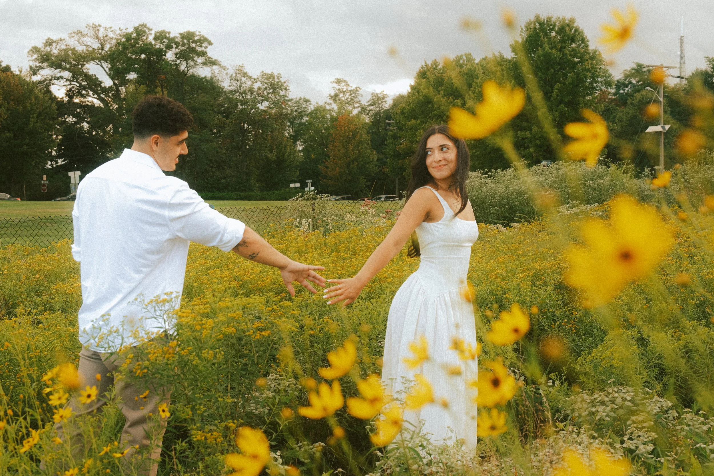 A couple holding hands and smiling in a yellow wildflower field with trees and cloudy sky in the background.