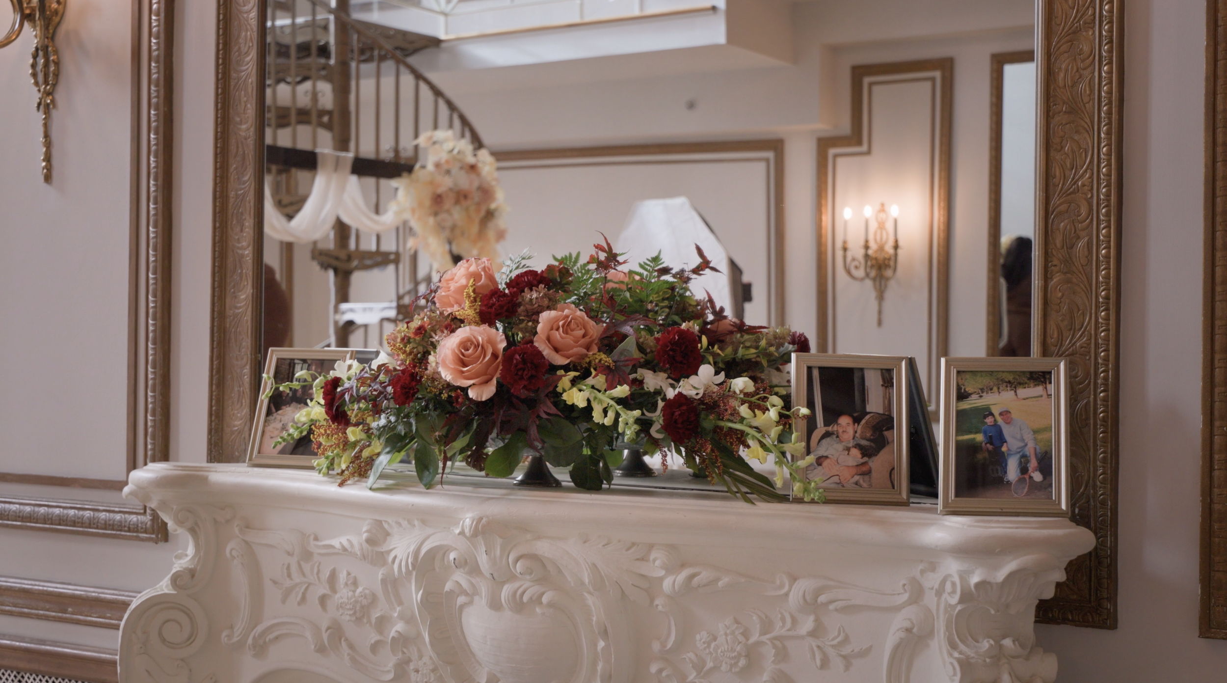 A white ornate fireplace mantel with a large floral arrangement of pink and red roses, white orchids, and various greenery, flanked by framed family photos, in a room with decorative wall moldings and sconces.