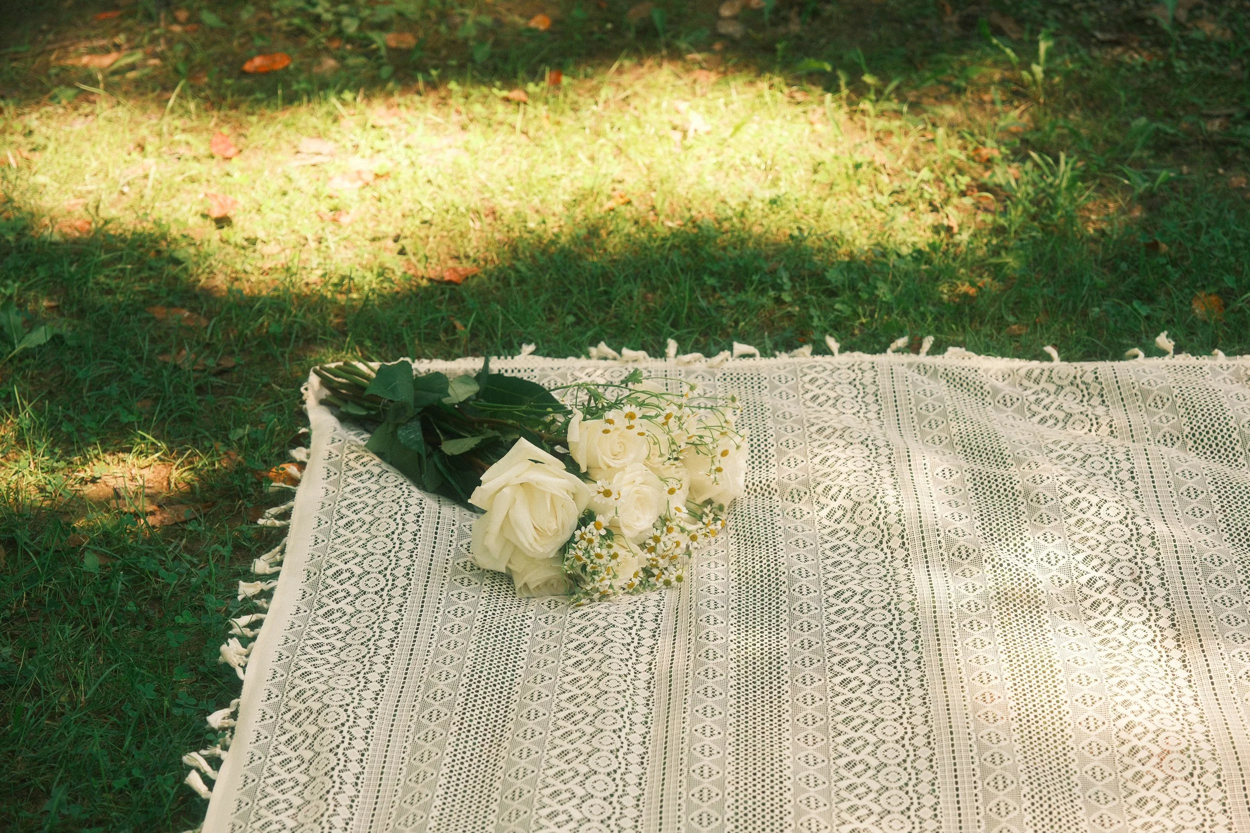 A bouquet of white roses and small white flowers rests on a white lace tablecloth outdoors on grass and dirt.