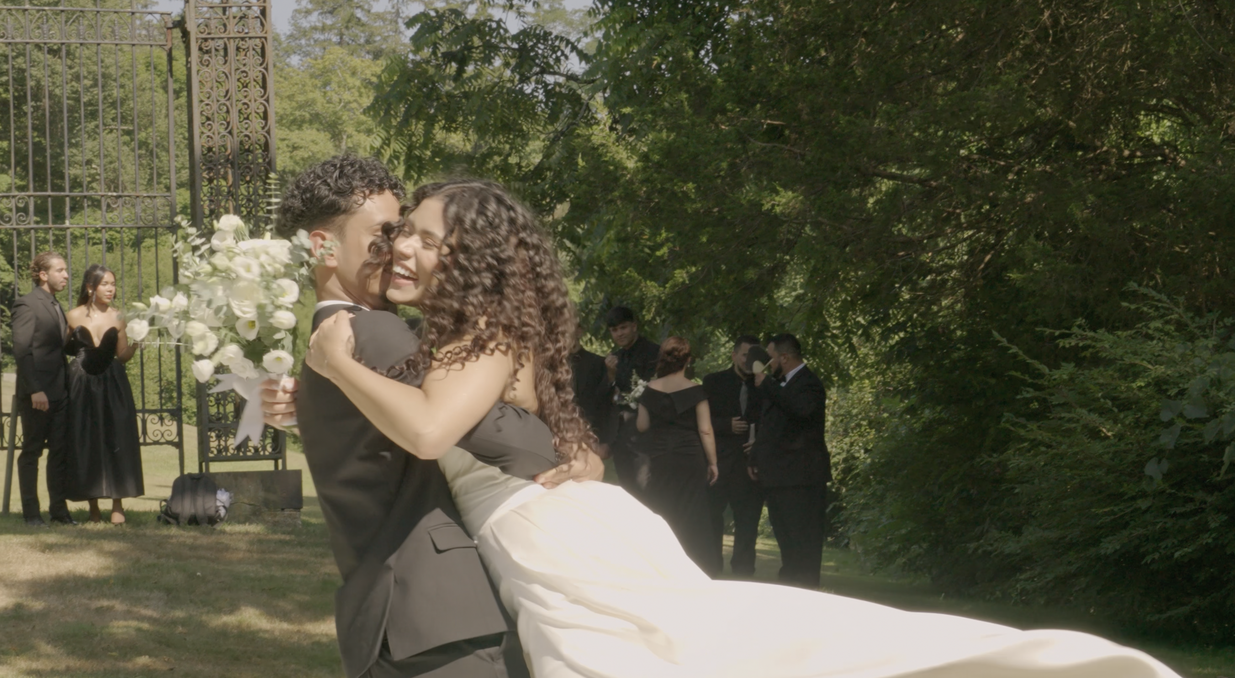 A joyful couple in wedding attire dancing and embracing at an outdoor wedding, with other guests in formal wear in the background.