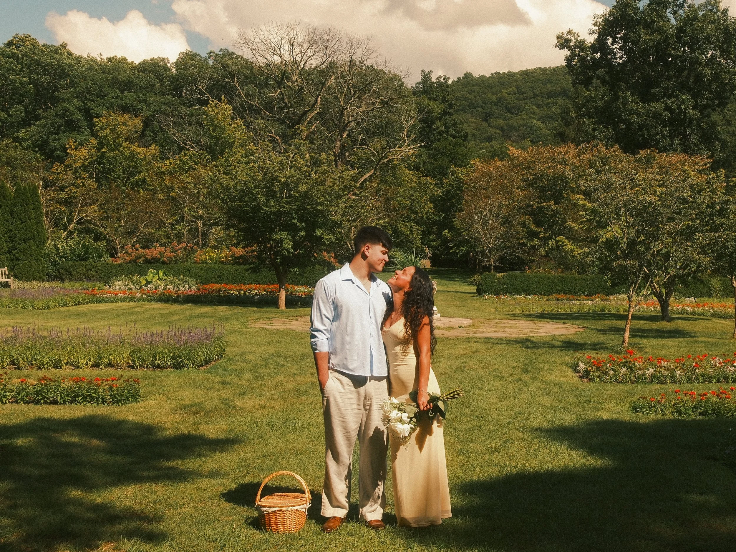 A young man and woman standing in a garden, gazing at each other lovingly. The woman is holding a bouquet of white roses and wearing a beige dress; the man is wearing a light-colored shirt and beige pants. There is a woven basket on the ground beside them. The background features a lush green landscape with trees, colorful flowers, and a partly cloudy sky.