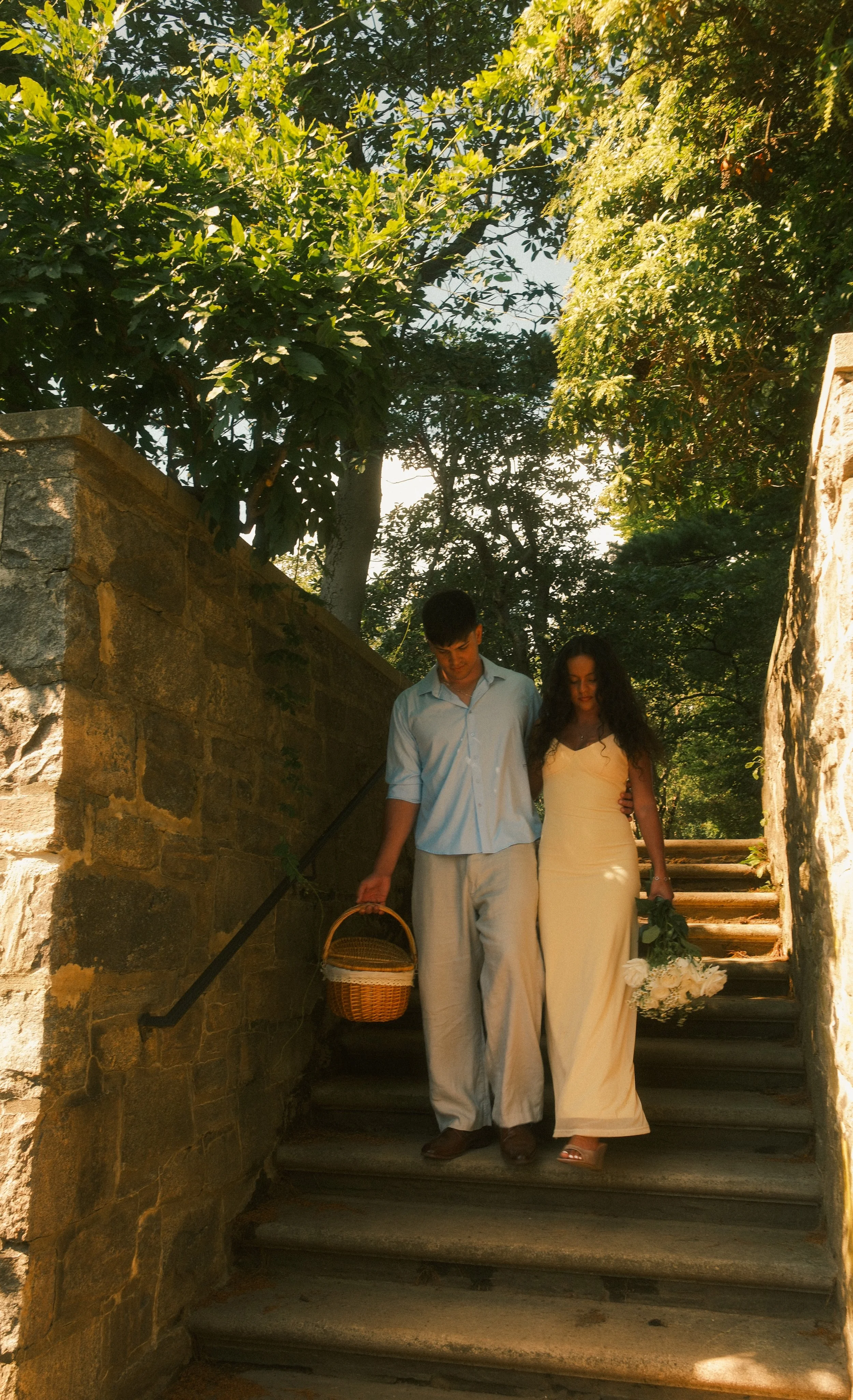 A couple walking down outdoor stone stairs, the man carrying a basket and the woman holding a bouquet of white flowers, surrounded by lush green trees and sunlight.