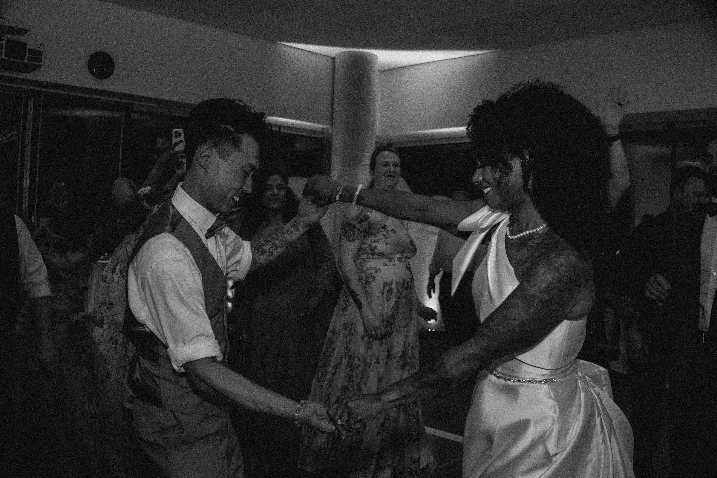 A couple is dancing peacefully in a dimly lit room at a celebration, with both smiling and holding hands.