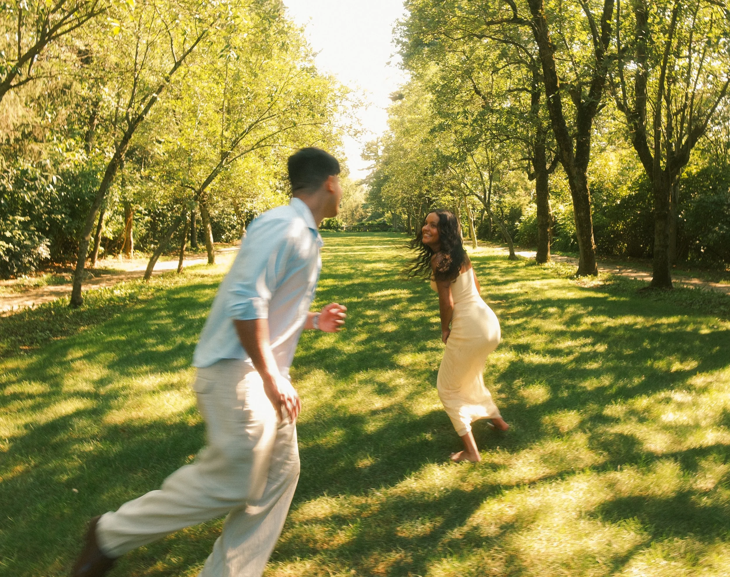 A man and a woman joyfully running and smiling in a park with green grass and trees during late afternoon.