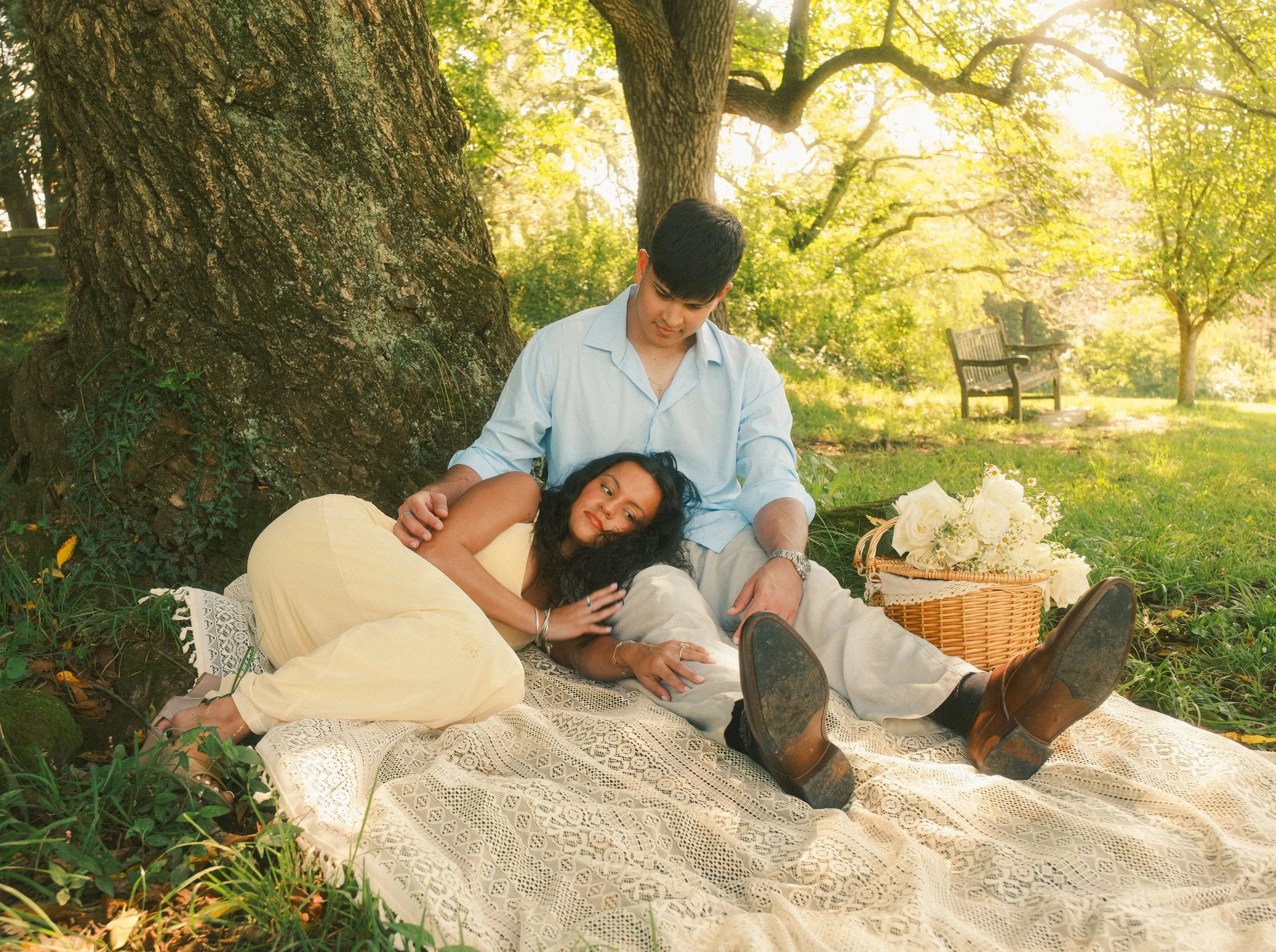 A young man and woman sitting outdoors under a tree on a blanket, surrounded by greenery, during daytime.