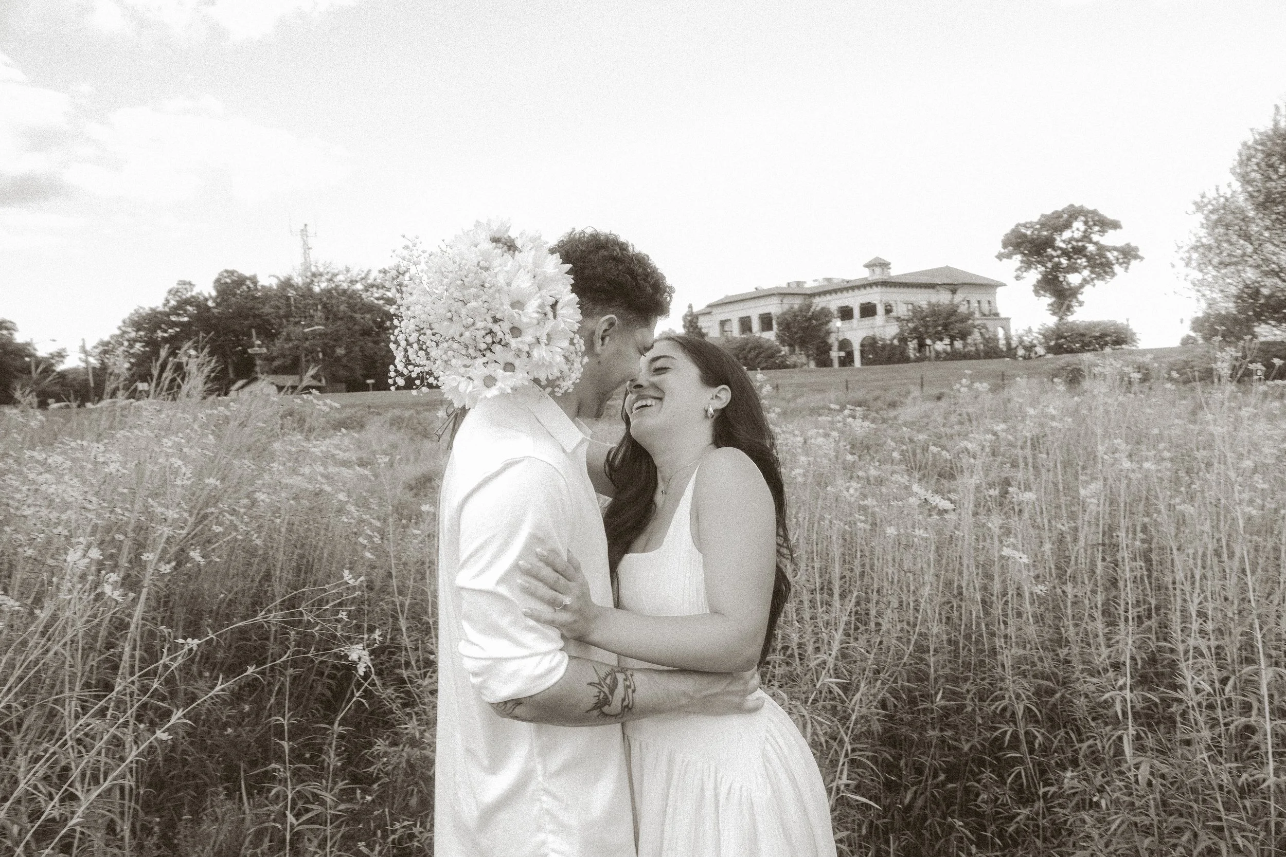 A couple standing close together in a grassy field, smiling and looking at each other, with a large house in the background. The man is wearing a white shirt and has a floral arrangement on his shoulder, and the woman is wearing a white dress.