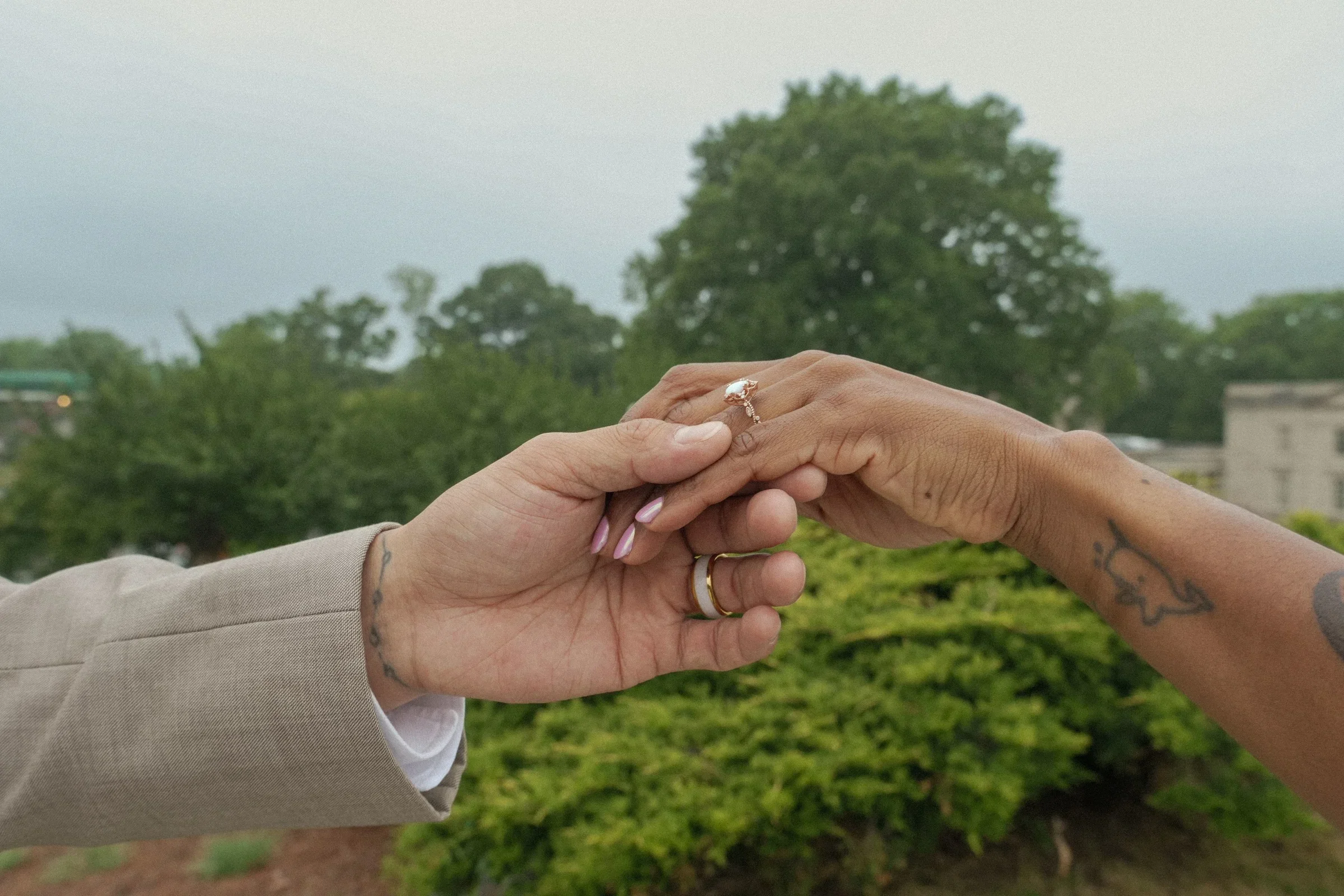 Two hands are holding each other outdoors with a background of trees and a cloudy sky. One person is wearing a beige suit; the other has tattoos on their forearm and wears rings, including an engagement ring.