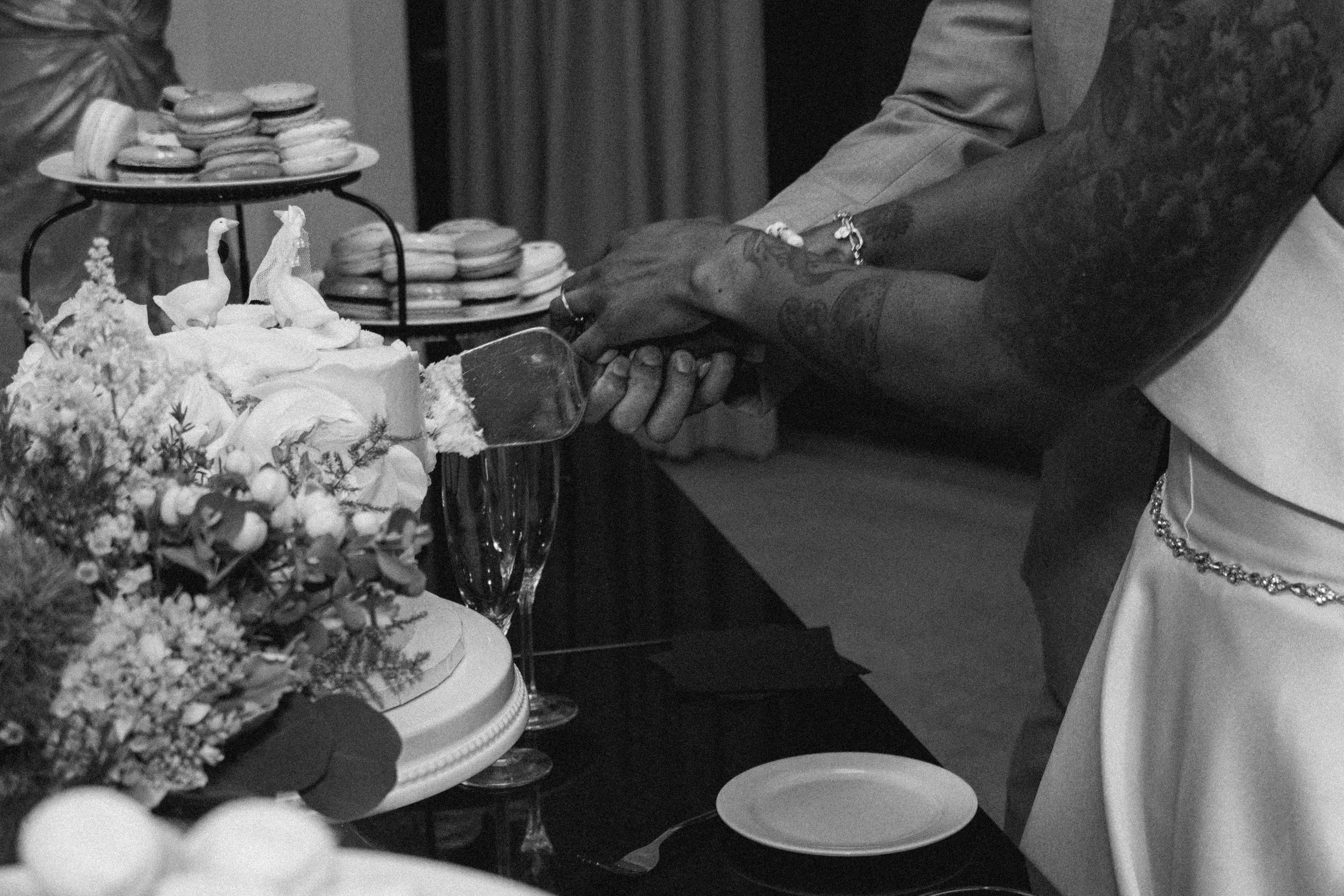 Two people holding hands next to a table with a cake, flowers, and macarons, at what appears to be a celebration or wedding.