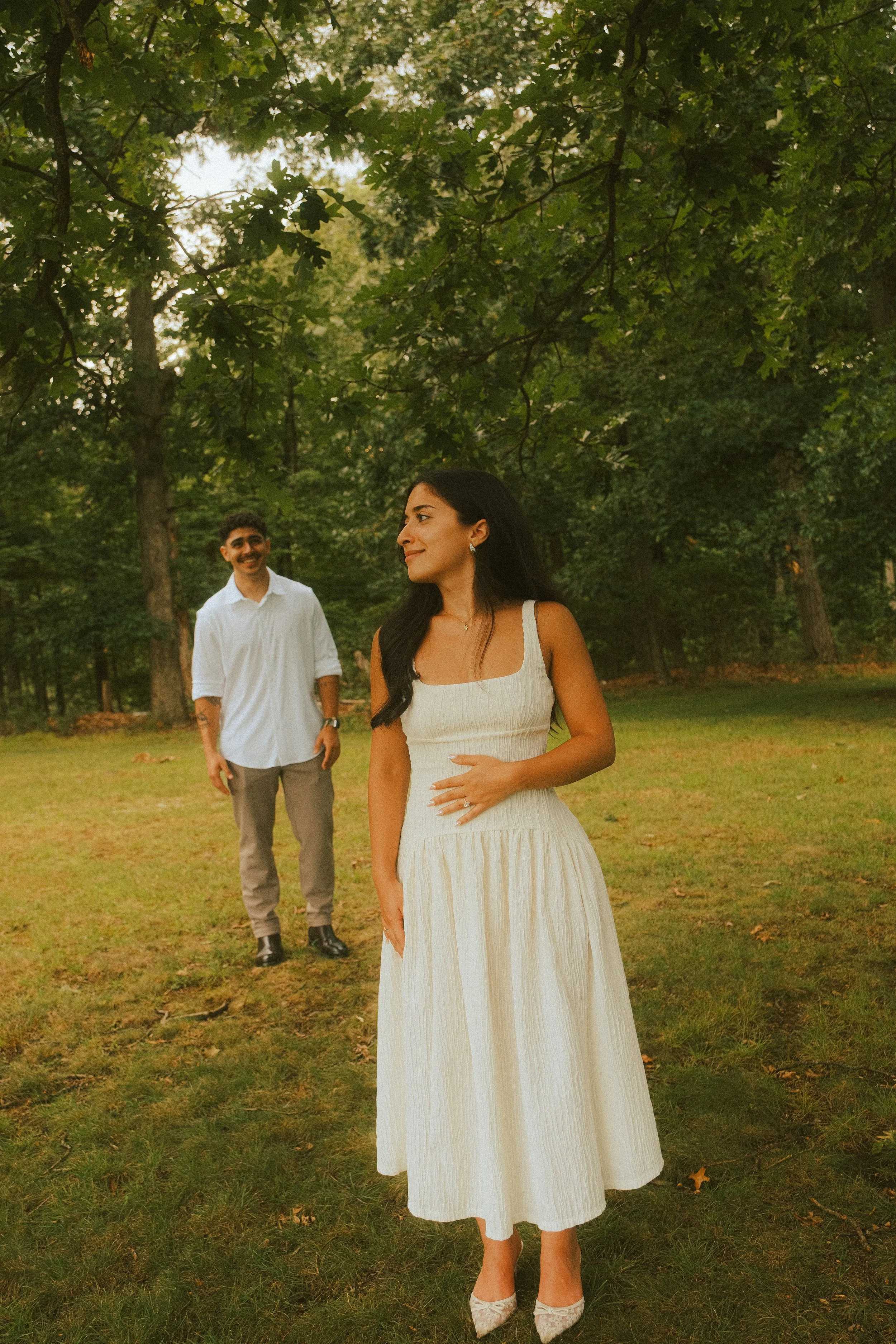 A woman in a white dress standing on a grassy area under trees, with a man in a white shirt and gray pants smiling in the background.