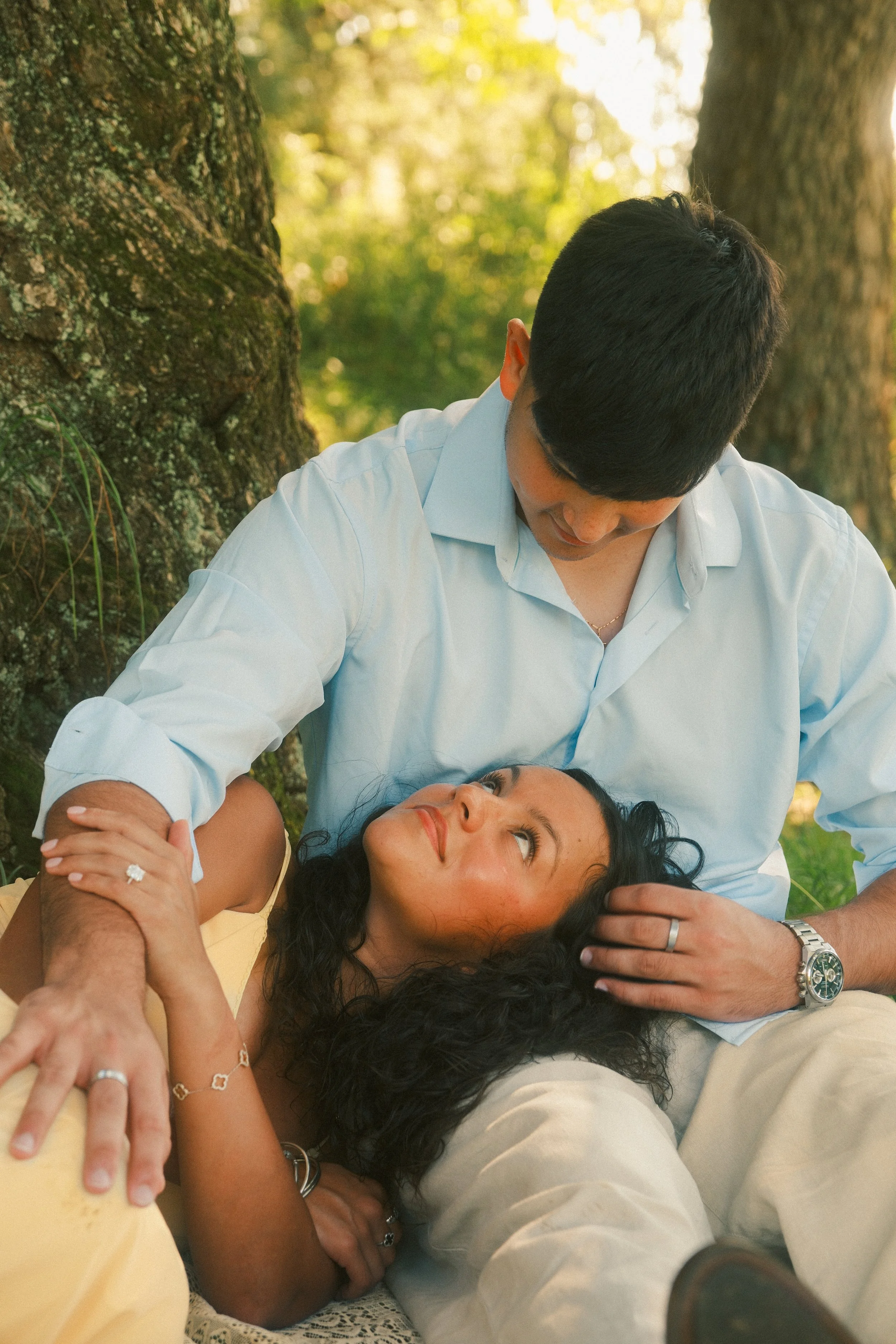 A man and a woman are lying down outdoors, with the man leaning over the woman as she looks up at him. They are surrounded by trees, and the scene appears to be set in a park or forested area during daylight.