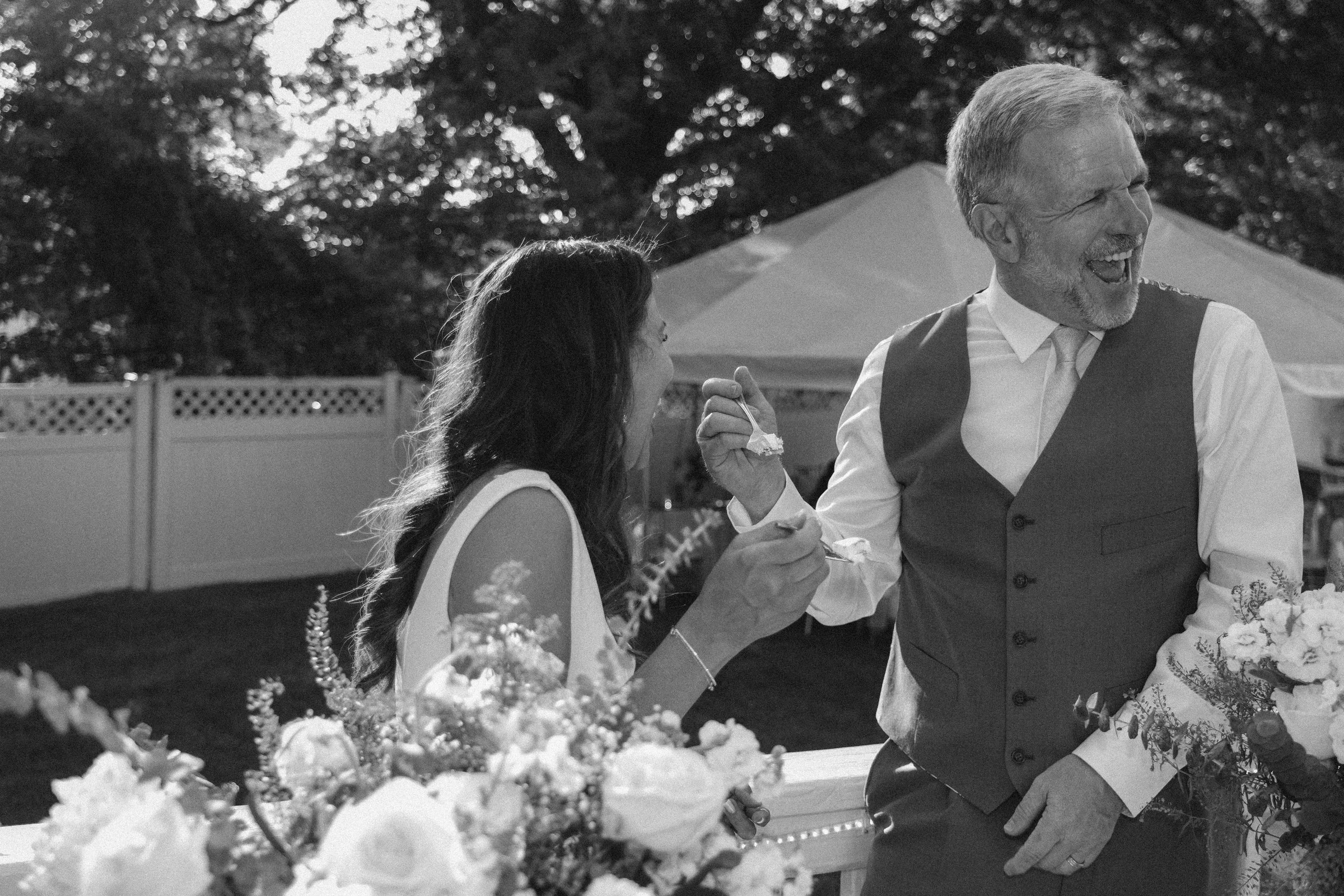 A bride and groom at a wedding reception outdoors, sharing a joyful moment with the groom holding a fork of cake and the bride reaching for it, decorated with flowers and a background of trees and a fence.