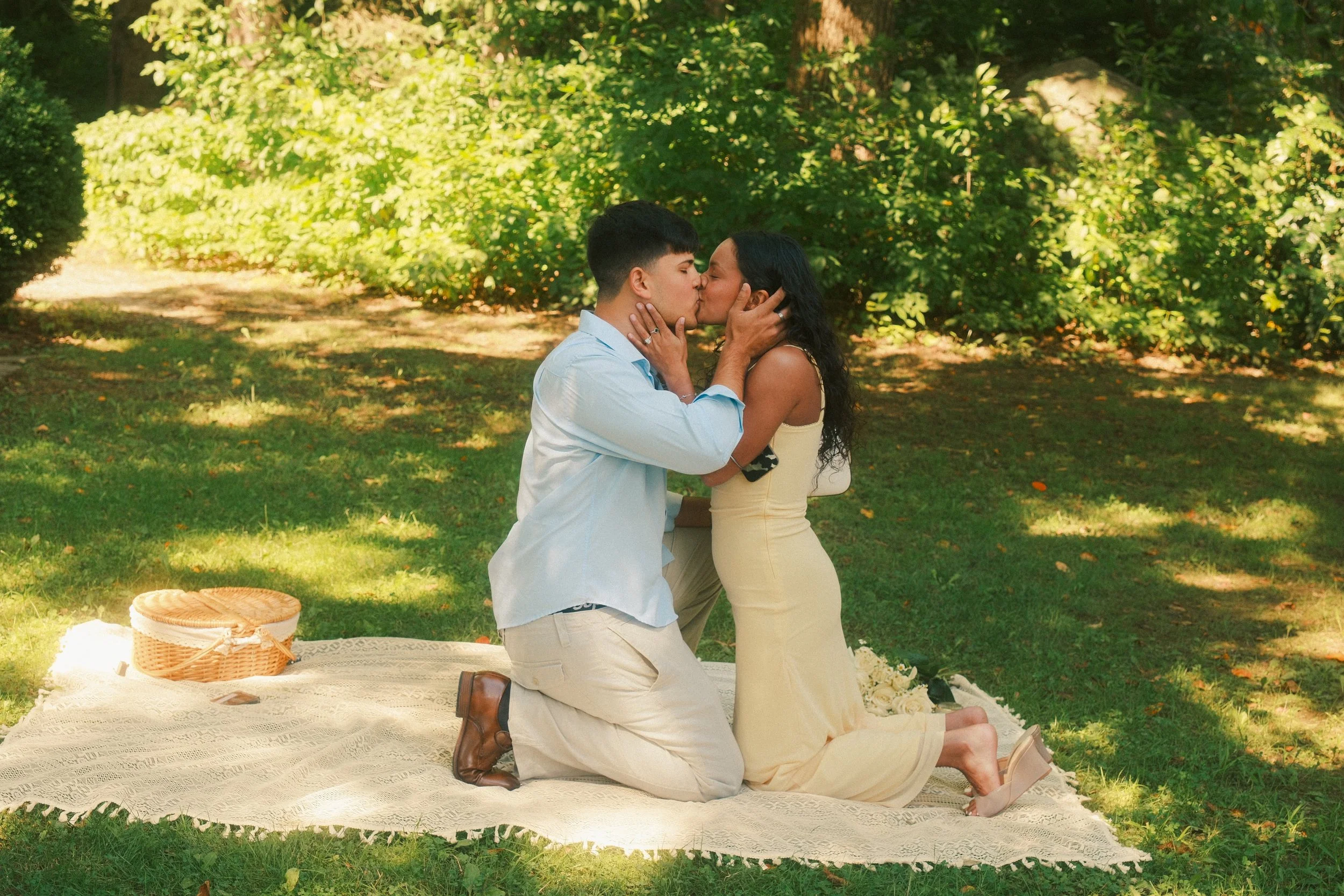 A couple is kneeling and kissing on a white lace blanket in a park with green trees. The man is wearing a light blue shirt and beige pants, and the woman is wearing a yellow dress. There are baskets and flowers on the blanket.