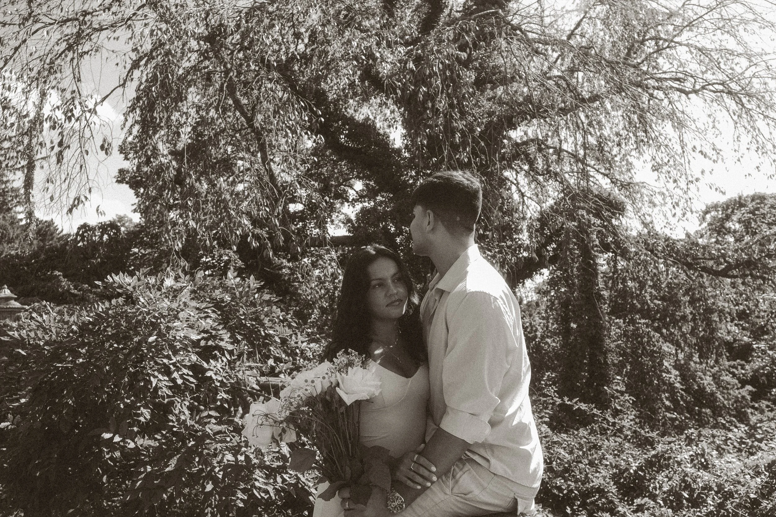 A couple standing close together outdoors surrounded by trees, with the woman holding a bouquet and looking at the man who is taller and facing her.