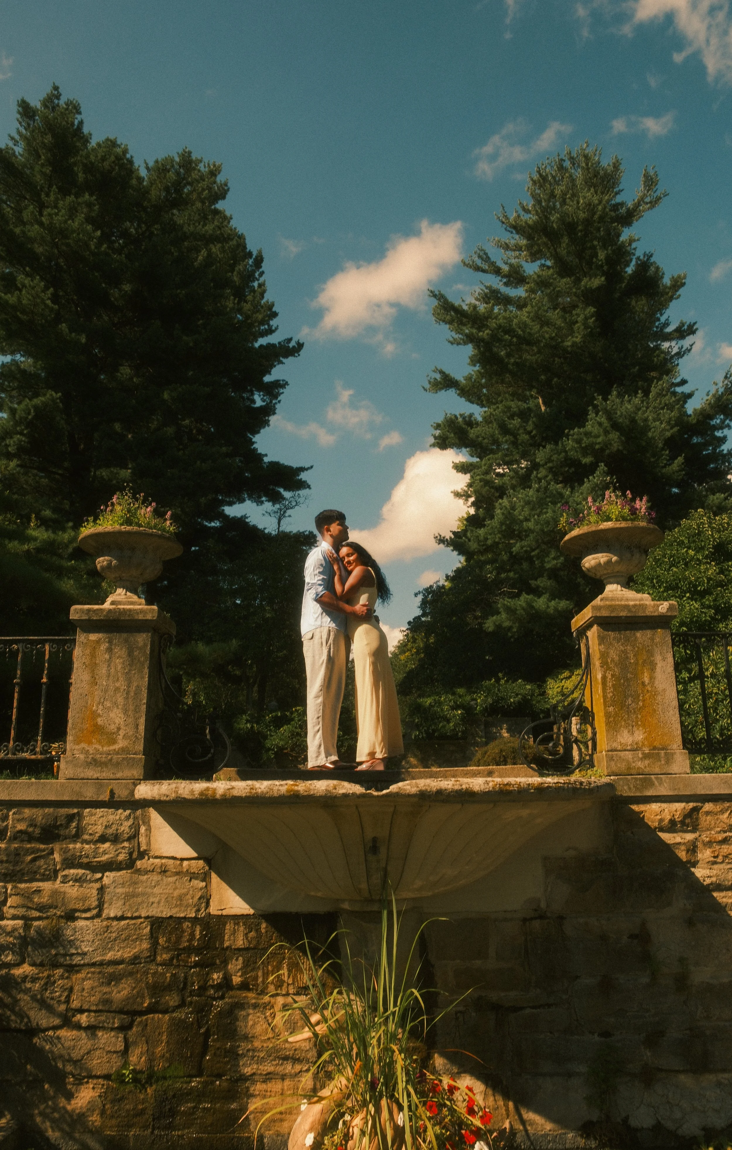 A couple standing close on a stone bridge surrounded by trees, with a blue sky and white clouds overhead, during daytime.