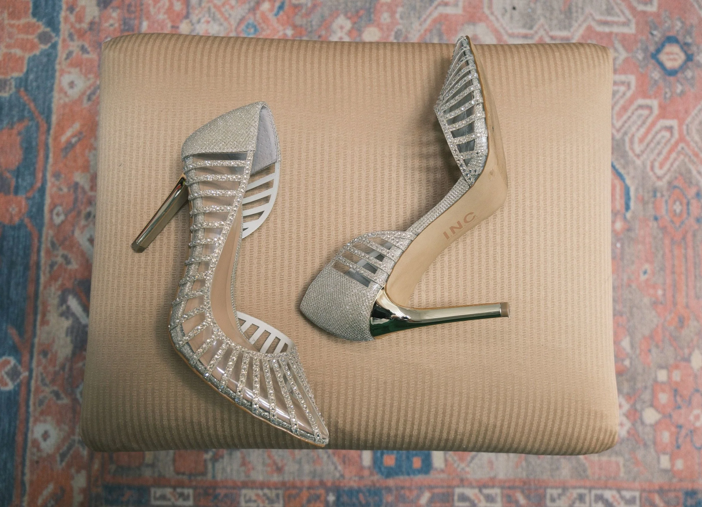 A pair of high-heeled shoes with silver glitter and open-toe design, placed on a beige textured surface with a pink and blue patterned rug underneath.