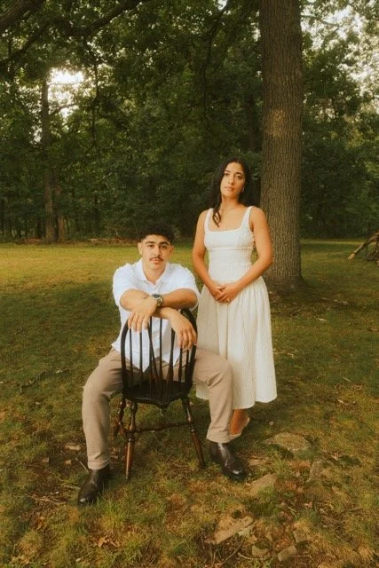 A man sitting on a black chair and a woman standing beside him in a park with trees, both dressed in white dresses, during sunset.
