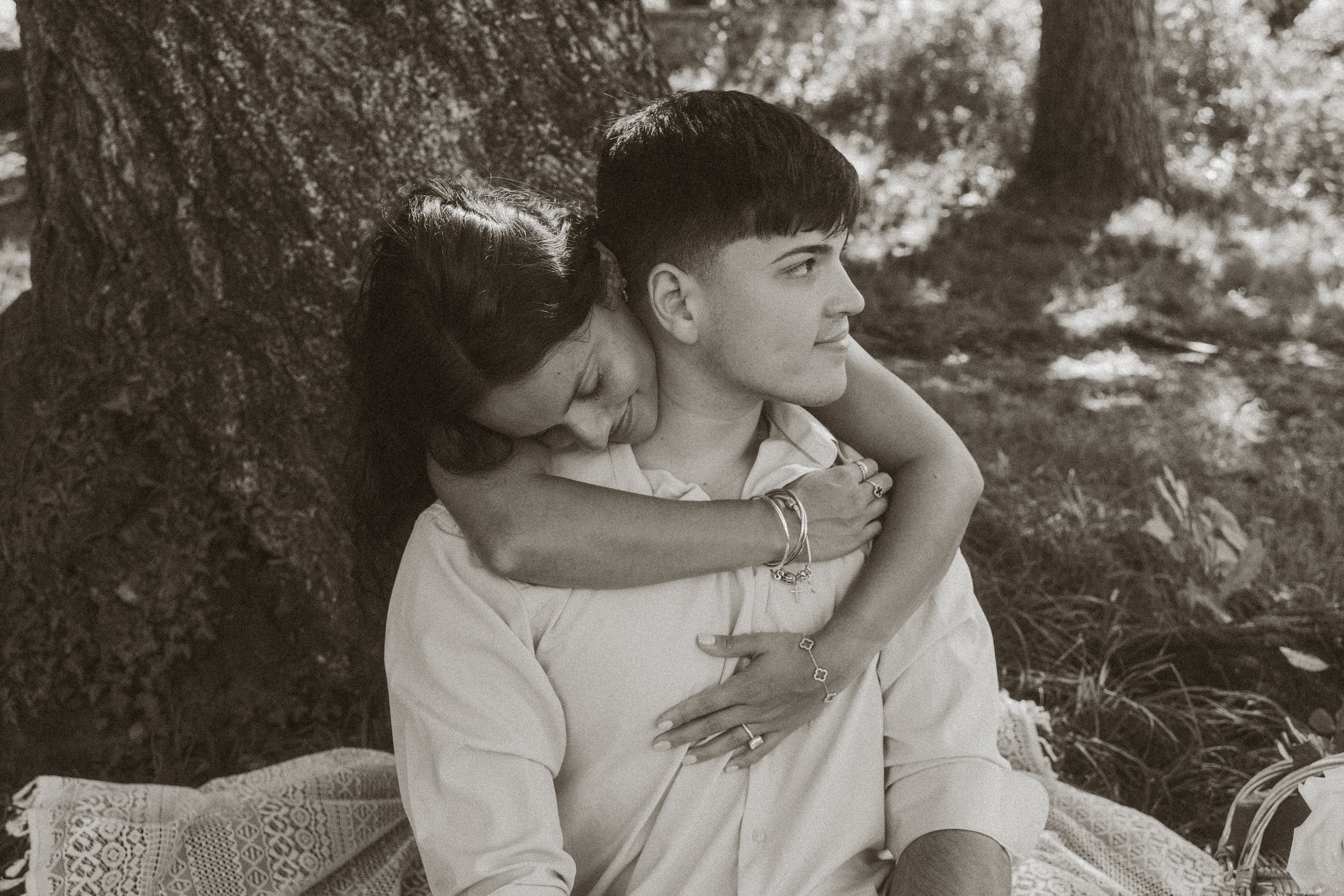 A young woman with short dark hair embraces a young man with short dark hair and a slight smile, sitting under a large tree in a park, both dressed casually in light-colored clothing.