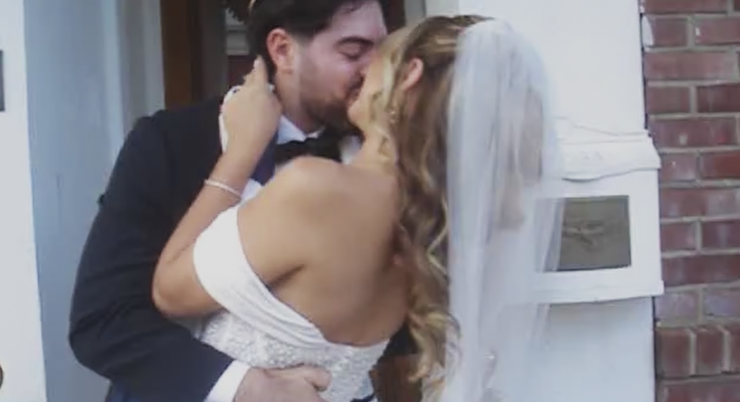 A bride and groom kiss outside a building, with the bride in a white dress and veil, and the groom in a tuxedo.